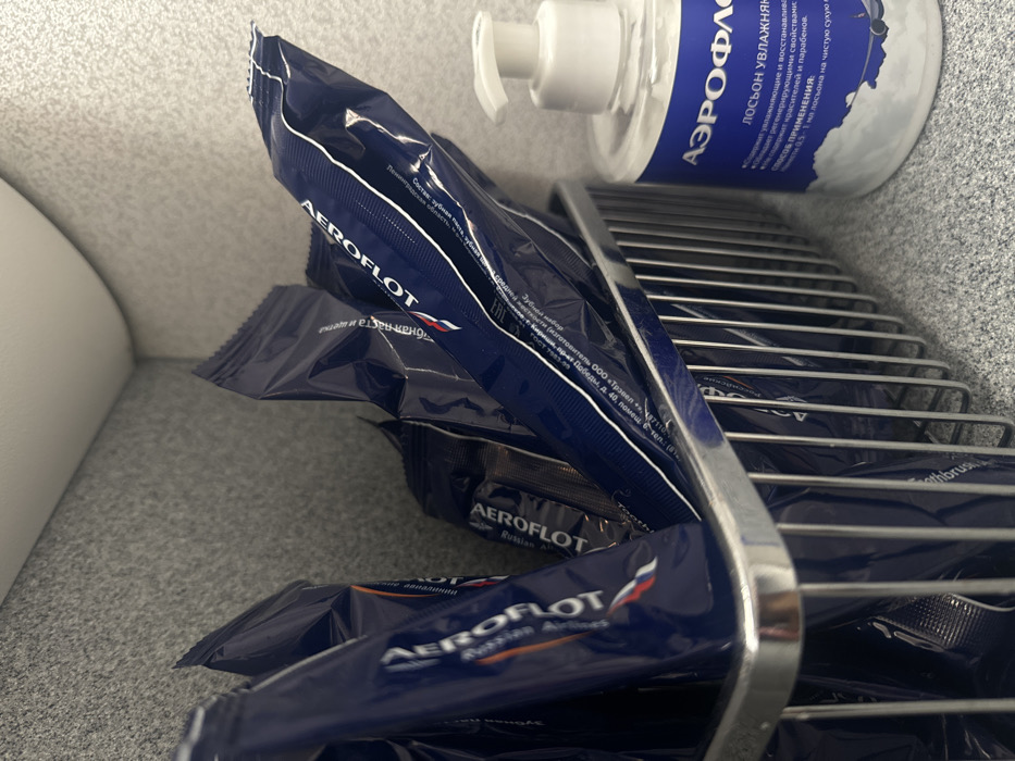The image showcases several Aeroflot-branded toothbrush and toothpaste packets arranged in a chrome wire rack. A bottle of Aeroflot lotion is also visible in the top right. The packets are dark blue with white lettering. The lotion bottle is white with blue text, stating that it is a moisturizing lotion with regenerating properties. The packets contain a toothbrush and toothpaste. The composition details the toothpaste's ingredients and manufacturing.