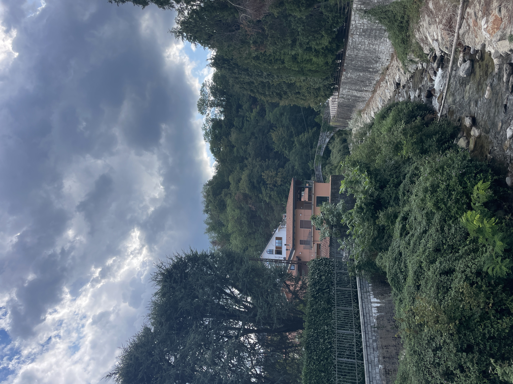 The image depicts a scenic view in the city of Pella, Italy, featuring a narrow, rocky stream flowing through a lush green valley. The stream, with clear water cascading over stones, is bordered by dense vegetation and a stone retaining wall on the right side. On the left bank of the stream, a metal fence runs along a path, with a wall behind it. Further back, a multi-story building with a terracotta roof is visible, nestled among trees and a hillside. A small arched bridge spans the stream in the middle ground, leading to a wooded area. The sky above is overcast with dramatic clouds, suggesting a possibly cloudy or stormy day. The overall scene conveys a tranquil, natural environment with human habitation integrated into the landscape.