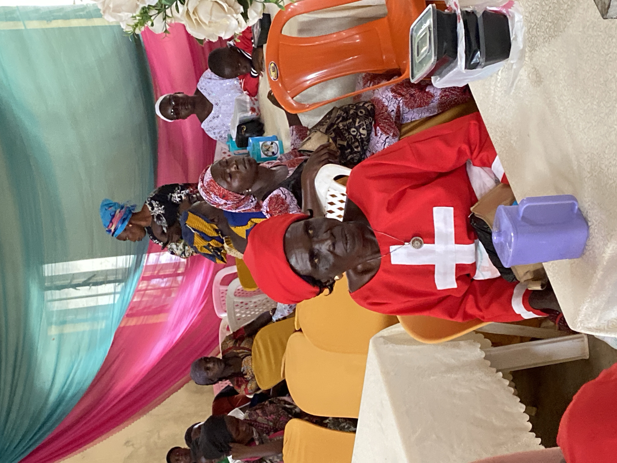 The image depicts an indoor gathering of people, likely a community event or service, in Riyom, Nigeria. In the foreground, an elderly woman wears a red headscarf and a red garment adorned with a white cross. She is seated at a table covered with a white tablecloth, with a purple mug and black takeout containers on it. Behind her, other individuals are visible, some wearing headscarves and patterned clothing. The background is decorated with draped fabric in shades of pink and teal, suggesting a festive or ceremonial occasion. Several chairs, some orange and some white plastic, are arranged around tables. The lighting appears to be natural, possibly from windows obscured by the fabric.