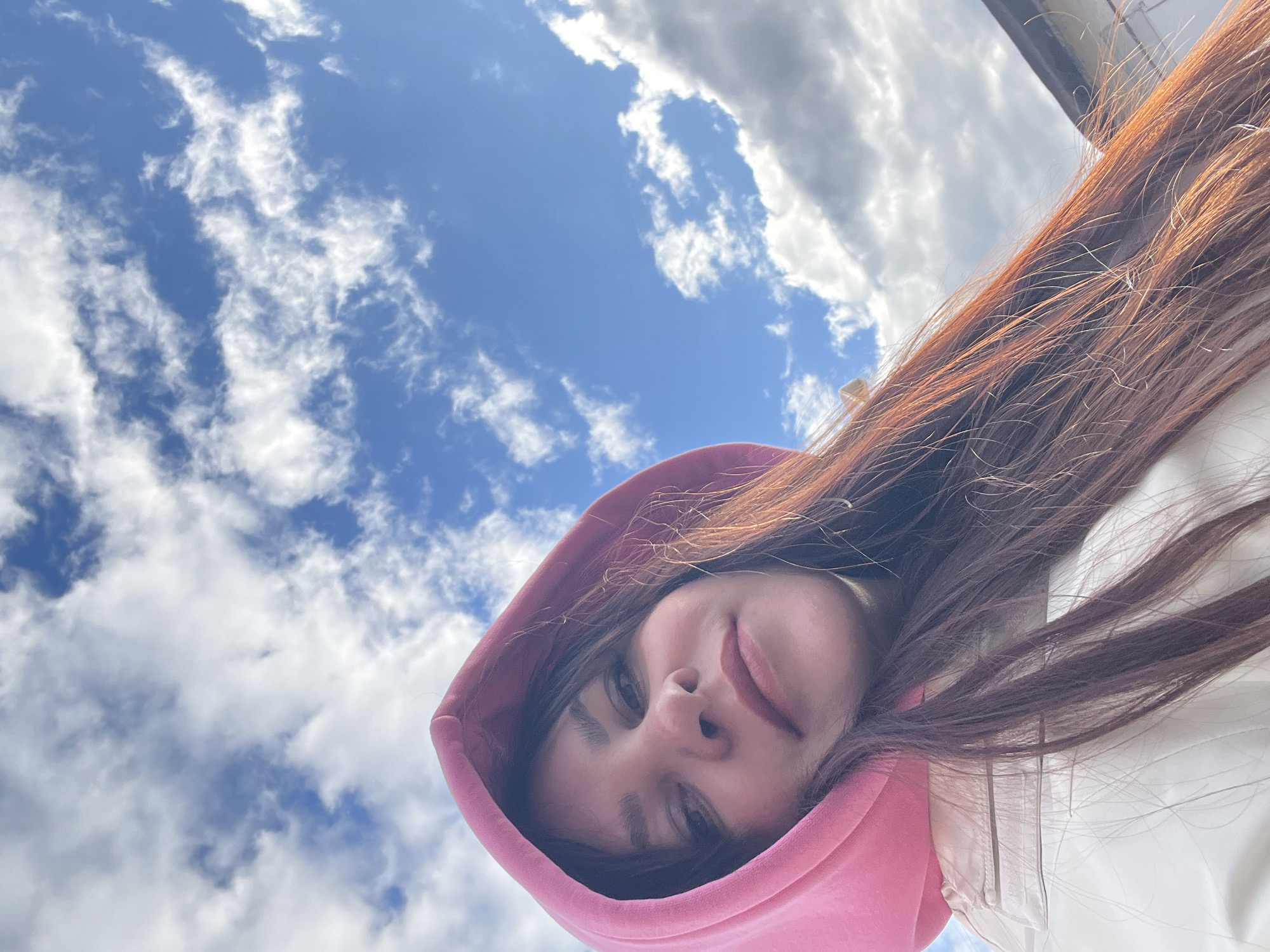 A low-angle close-up shot shows a woman's face framed by a pink hoodie. She looks directly at the camera with a slight smile. Her long brown hair cascades toward the top right corner. The background is a bright blue sky with scattered white clouds. Part of a building is visible at the top right. There is nothing to confirm the city name provided.