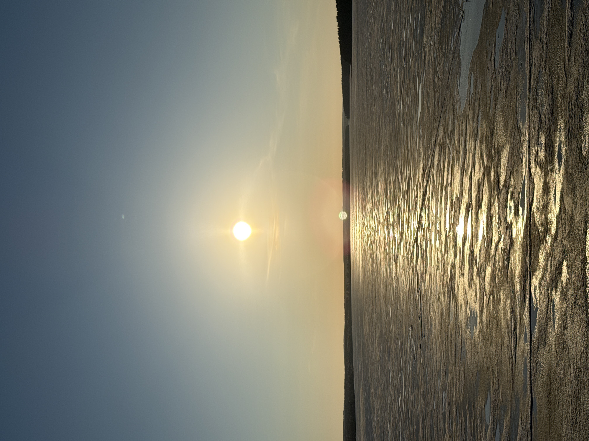The image depicts a sunlit landscape with a body of water in the foreground and a clear sky above. The sun is positioned in the center of the frame. The water surface reflects the sunlight, creating a shimmering effect. A dark treeline can be seen along the edge of the water.
While Votkinsk, Russia is known for its industrial history, the image does not contain any elements that would indicate its location in that specific city. There are no distinct landmarks visible that would allow for verification.