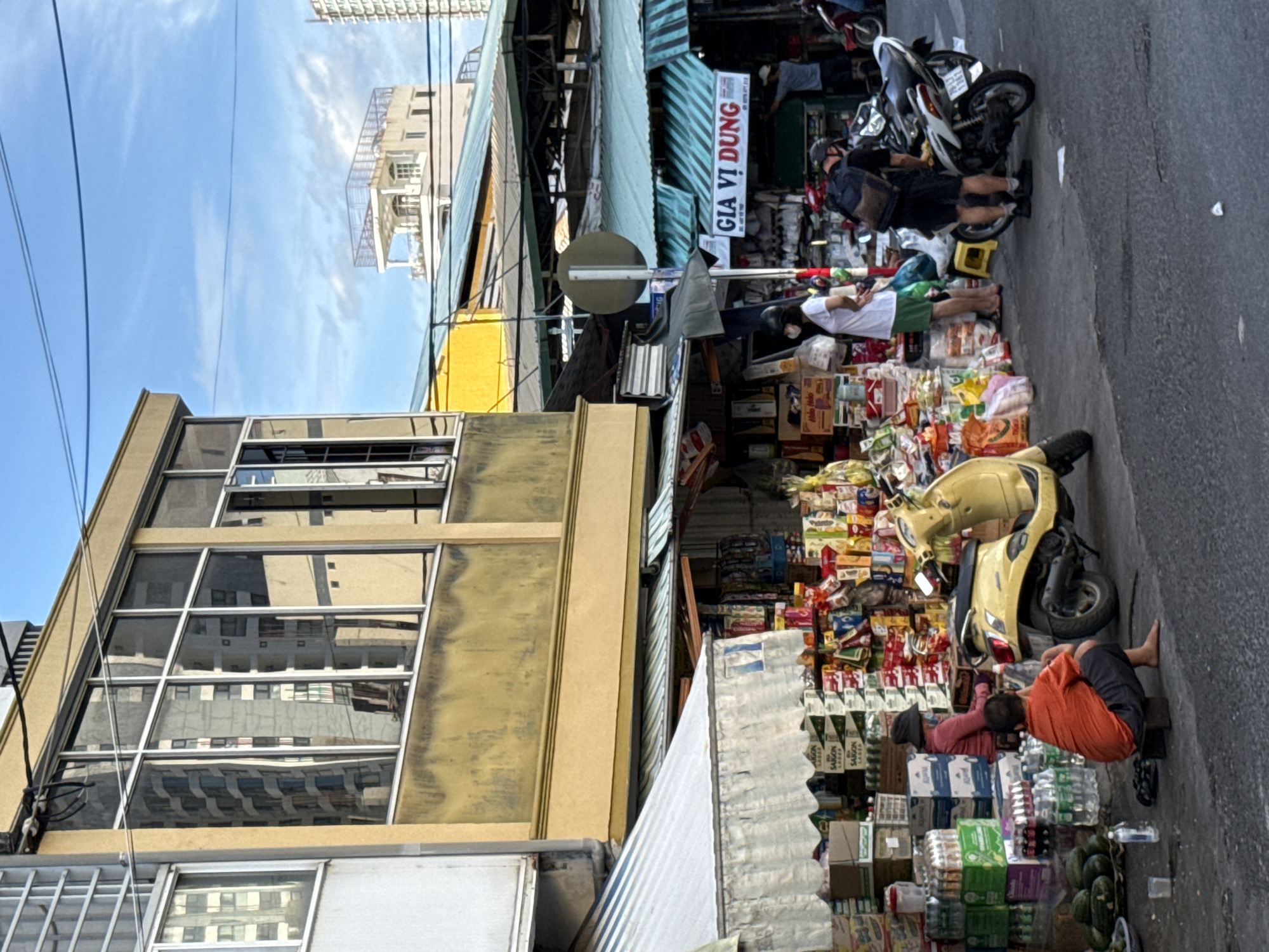 This image depicts a street market scene in Nha Trang, Vietnam. The market is situated in front of a multi-story building with a yellow facade and large windows reflecting the surrounding environment. Various stalls are set up along the street, covered by awnings made of striped fabric. These stalls display a wide assortment of goods, including bottled drinks, packaged food items, and other merchandise.
In the foreground, two individuals are present. One, wearing an orange shirt and dark shorts, is sitting on a small stool on the ground, appearing to be resting or observing the surroundings. Next to them are several large green vegetables, possibly gourds or cucumbers, arranged on the pavement. A yellow scooter is parked behind this individual.
Further into the scene, several other people are visible. A woman in a pink top and dark hat is seated near a stack of boxes. Another person, wearing a white t-shirt and green shorts, is standing near a stall, interacting with the market. Two men are standing by motorcycles parked on the street. One man, wearing a black backpack, is leaning over a white motorcycle. Another individual, wearing a grey t-shirt and dark shorts, is standing near a stall in the background.
The environment shows a typical urban street with asphalt pavement. Overhead, power lines are strung across the sky, and the sky itself is blue with scattered clouds. The architecture visible in the background suggests a dense urban setting with modern buildings. A sign with Vietnamese text, "GIA VI DUNG," is prominently displayed above one of the market stalls.
