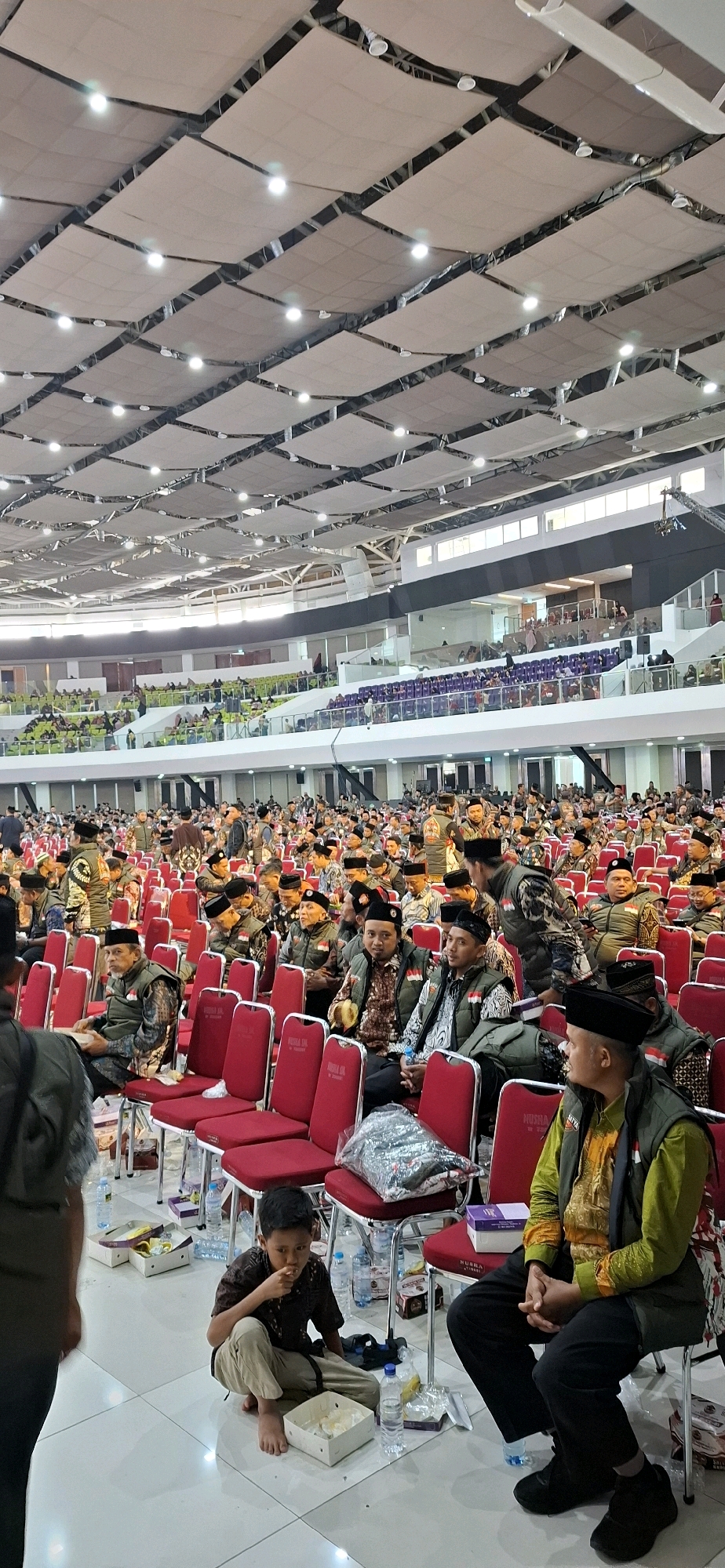 The image captures a large indoor gathering of people, likely a conference or religious event, in Blulukan, Indonesia. The venue is a spacious hall with tiered seating, characteristic of a convention center or stadium. The ceiling features a distinctive, modular design with fabric panels and integrated lighting. A vast crowd is visible, primarily composed of men, many wearing dark vests over patterned shirts and traditional caps, suggesting a shared affiliation or uniform. Some individuals are seated in rows of red chairs, while others are standing or moving through the aisles.
In the foreground, a young boy is sitting on the floor, eating from a white rectangular container. Bottles of water and other food items are scattered around him and other seated attendees. The atmosphere appears organized and orderly, with attendees focused on the event. The presence of multiple rows of identical red chairs and the sheer scale of the crowd indicate a significant gathering. The overall impression is one of a formal event with a large number of participants in a modern, well-equipped venue.