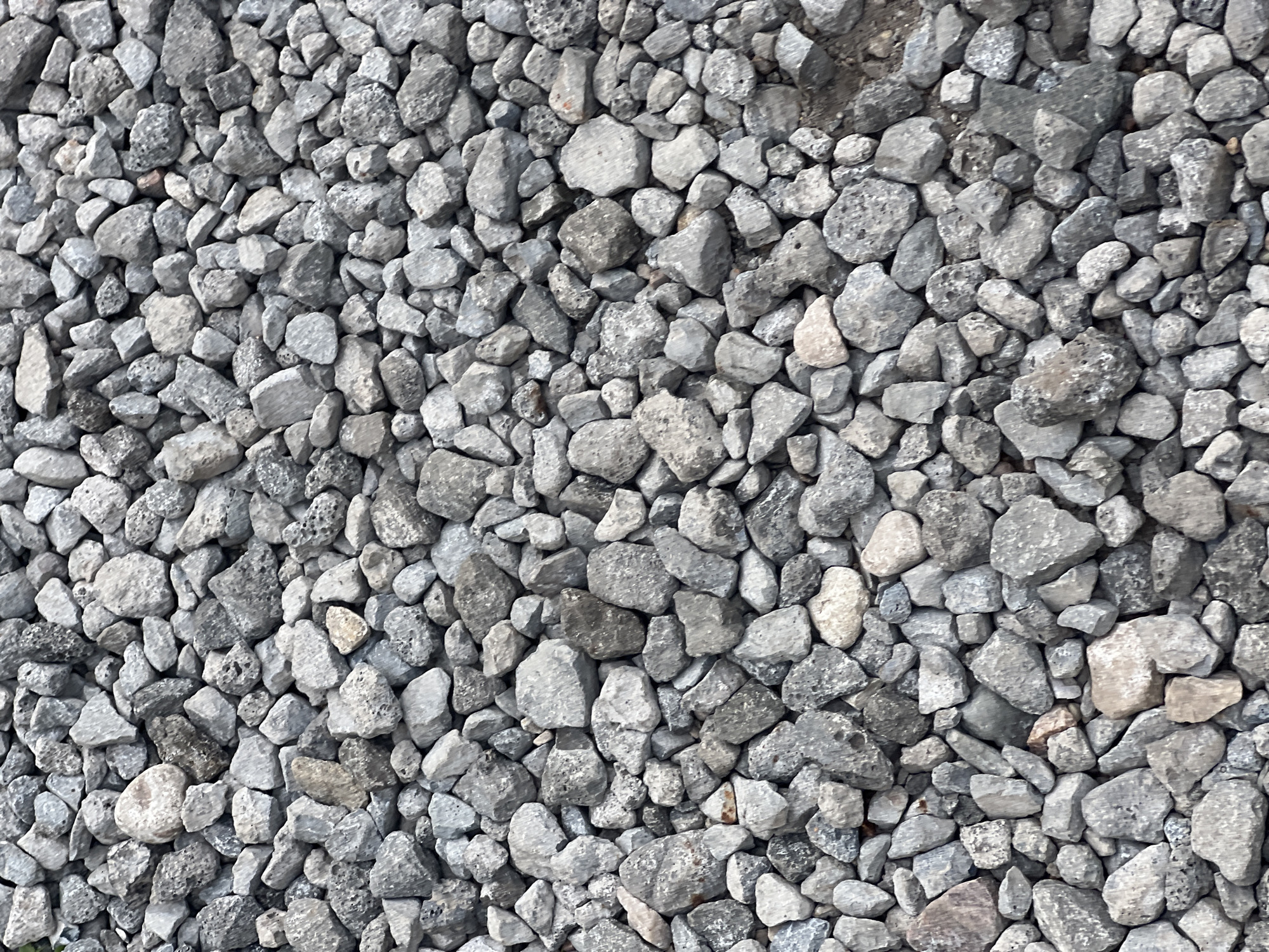 The image shows a dense, overhead view of a pile of rough, gray gravel. The gravel stones are of varying sizes and shapes, with sharp edges and irregular surfaces. They are a mix of light gray, dark gray, and some with hints of brown or white. The texture is predominantly rough and uneven. The lighting appears to be natural and diffused, casting no strong shadows, suggesting it might be overcast or the image was taken in indirect light. There are no discernible people, animals, or man-made objects other than the gravel itself. The environment appears to be an outdoor area, likely a construction site, pathway, or garden bed, common in urban or suburban settings within Unknown, Unknown.