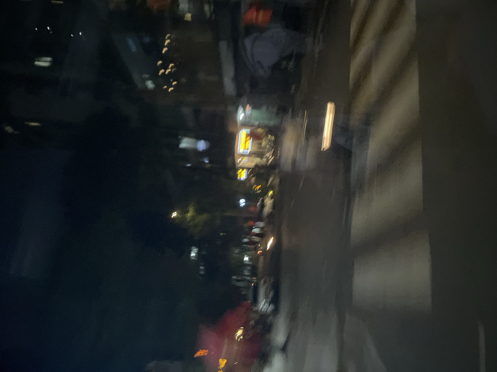 The image depicts a blurry night scene on a city street in Thủ Đức, Vietnam. A white car is visible in the center of the frame, appearing to be stationary or moving slowly. To the left, a red awning or umbrella suggests a roadside establishment. Further down the street, there are illuminated signs, likely from shops or restaurants, and the lights of buildings are visible in the background. A pedestrian crossing is marked on the road, with horizontal white stripes. The overall impression is a dimly lit urban environment at night, characterized by motion blur from the camera.