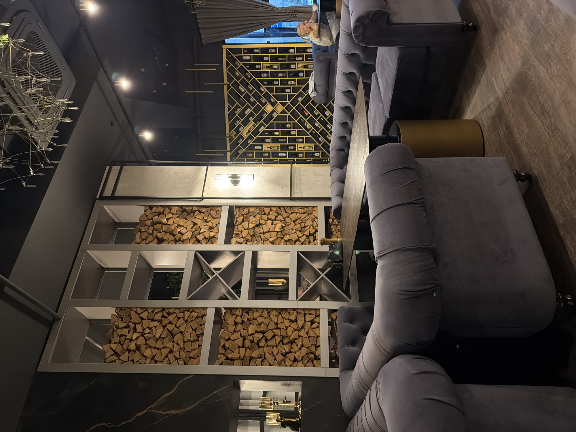 This appears to be an interior shot of a restaurant or bar. There is a display shelf filled with firewood. Beside the shelf is a long grey velvet sofa with tufted details. In front of the sofa is a dark tabletop and a cylindrical gold table. Behind the sofa is a display of wine bottles on a gold trellis. A person is sitting in front of the window and appears to be talking on a cell phone. The photo appears to be taken in Krasnoyarsk, Russia.