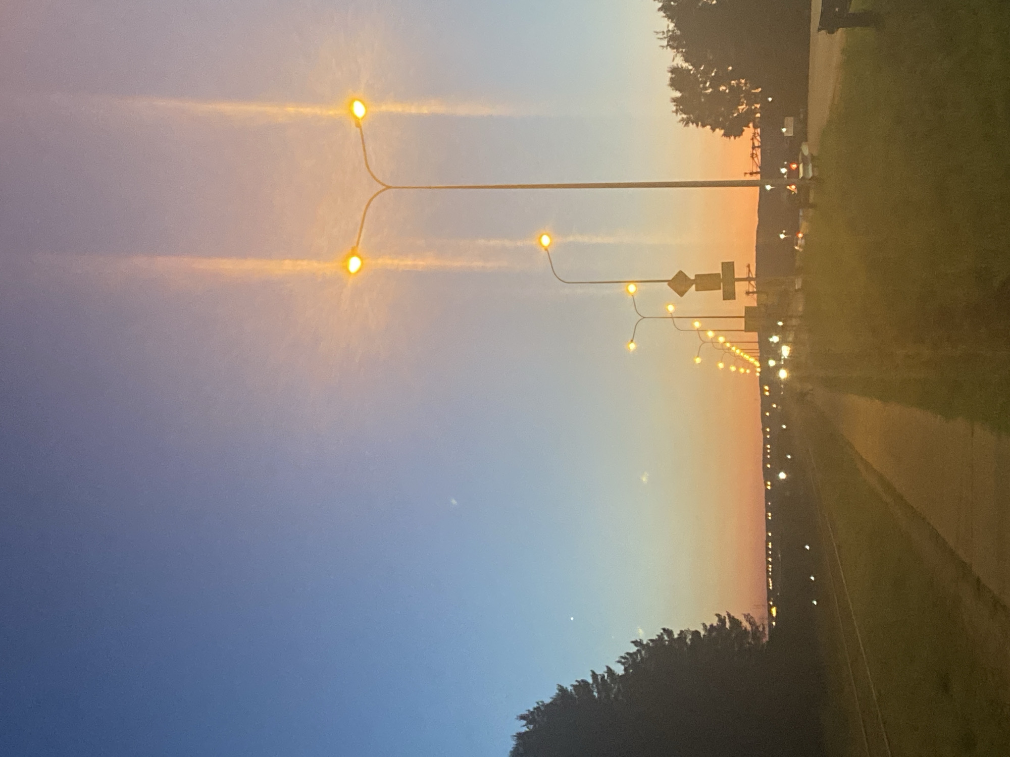 The image depicts a street scene at dusk. A row of streetlights illuminates a paved walkway next to a grassy area. The sky transitions from a deep blue at the top to orange and pink hues near the horizon, indicating sunset. In the background, more streetlights and distant lights suggest a populated area. Trees line the left side of the image, silhouetted against the twilight sky. The overall impression is one of an urban or suburban environment at the end of the day, with artificial light beginning to dominate the natural light.