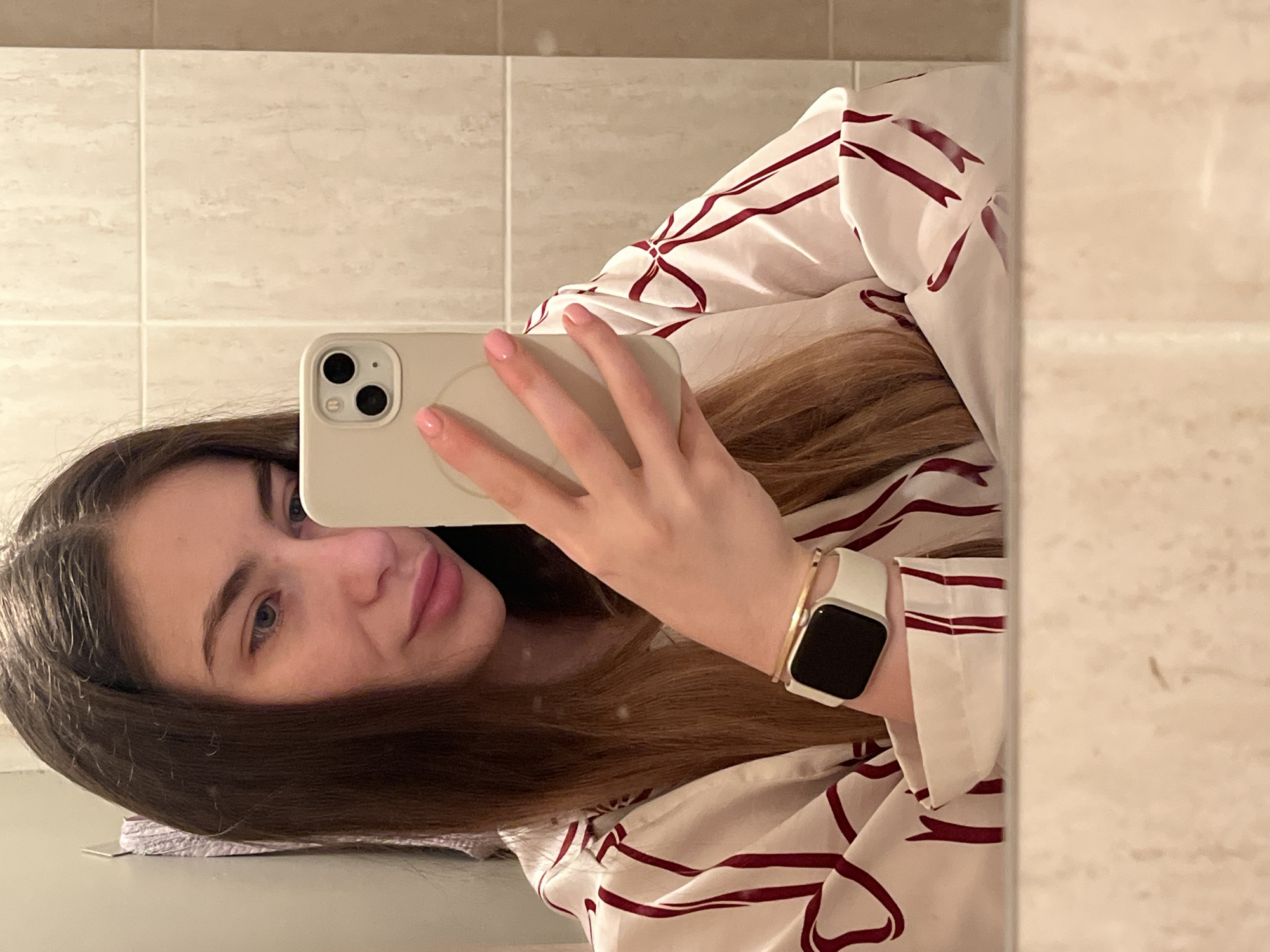 A woman is taking a selfie in what appears to be a bathroom. She is holding a light-colored smartphone up to her face, obscuring part of her features. Her hair is long and brown. She is wearing a white and maroon patterned shirt and a white smartwatch with a dark screen. The background consists of light-colored tiled walls and a mirror. The location is a bathroom in the city of Unknown, Unknown.