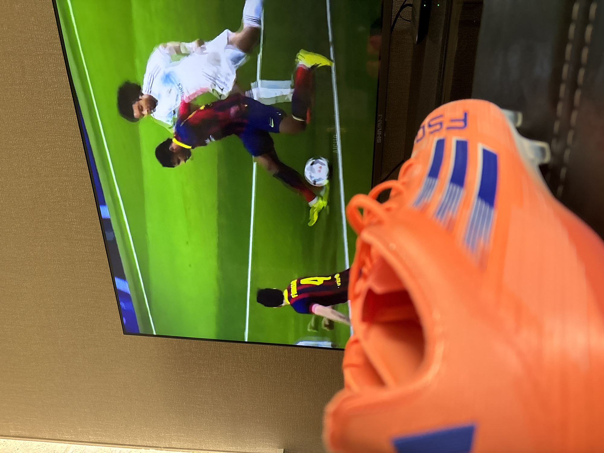 A still image depicts a television screen in the background displaying a soccer match, with a close-up of a bright orange soccer cleat in the foreground, partially obscuring the screen. The soccer cleat has blue stripes and the letters "F50" visible on the side. On the screen, two teams are competing on a green field, one in white uniforms and the other in blue and red. A soccer ball is in play near the player in the blue and red uniform. The environment appears to be a residential or living space with a textured wall in the background on the left. There is no indication of the city of Yerevan, Armenia.