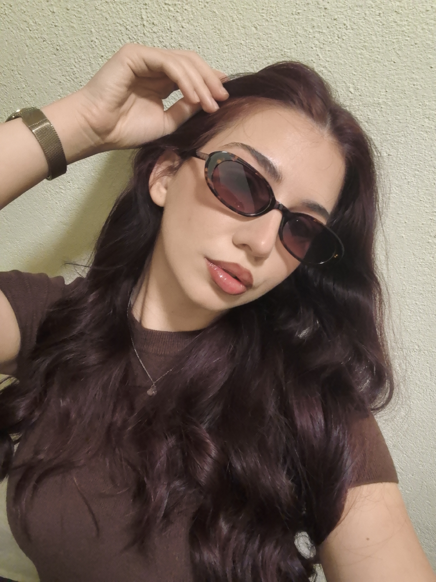 A young woman is standing in front of a light yellow wall. She is wearing a brown t-shirt and sunglasses, and has dark brown hair with purple highlights. She is wearing a brown watch on her left wrist and a silver necklace. She is touching her hair with her right hand. The location is Unknown, Unknown.