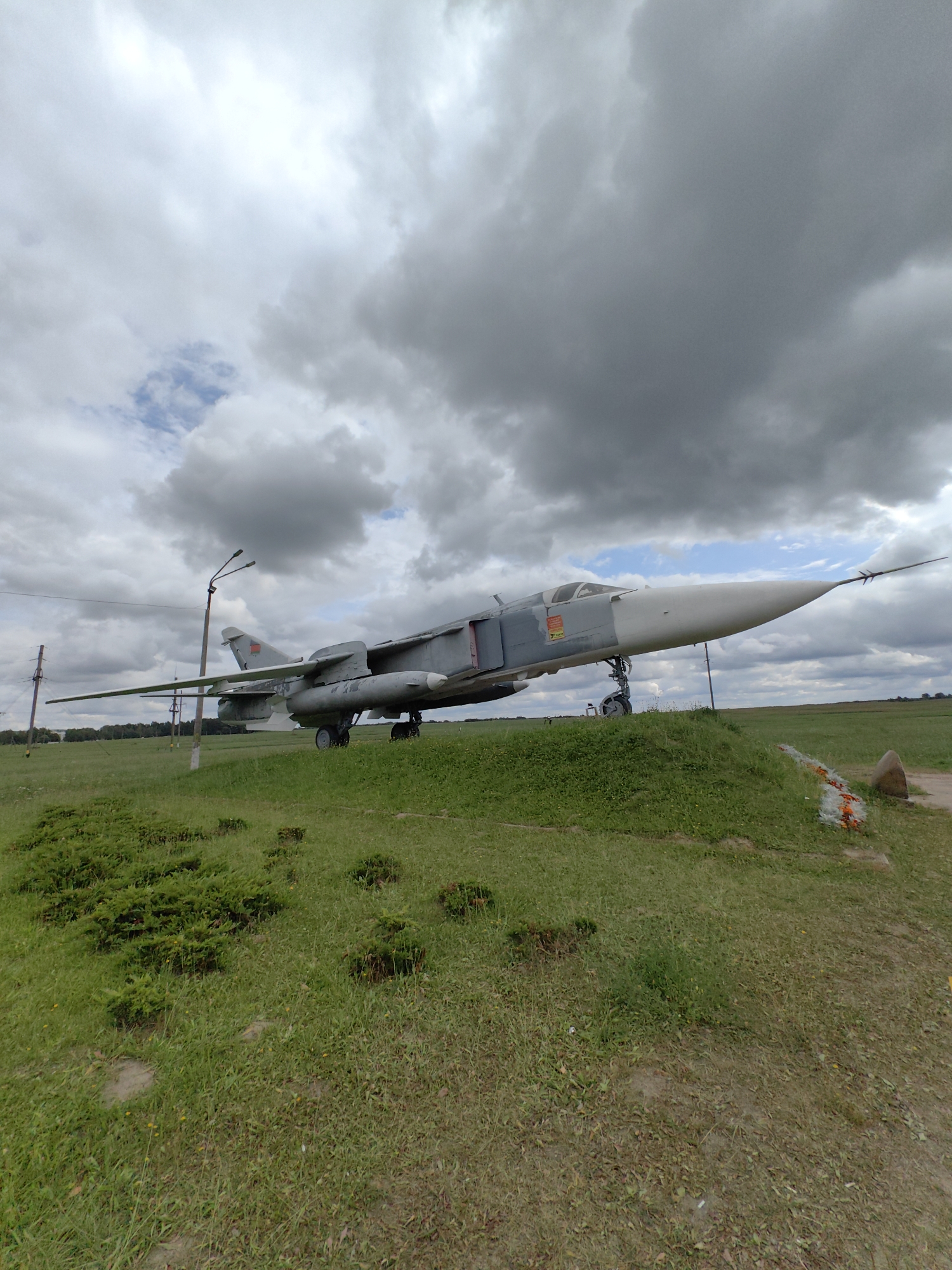 This is a low-angle, wide-shot, outdoor photograph of a Soviet-era jet fighter aircraft on display. The aircraft, a gray two-engine jet with swept wings and a distinctive nose cone, is positioned on a grassy mound. It is oriented facing towards the right of the frame. The foreground consists of a grassy field with some low-lying green bushes and scattered yellow wildflowers. In the background, beyond the aircraft, is a flat, open field with a treeline in the far distance. The sky above is filled with heavy, dark gray clouds, with a small patch of blue sky visible towards the top left. Two utility poles with lights are visible to the left of the aircraft, and a dirt path runs along the right edge of the frame. This scene is located in Pastavy, Belarus, likely at a museum or memorial site, given the static display of the aircraft. There are no people visible in the image.