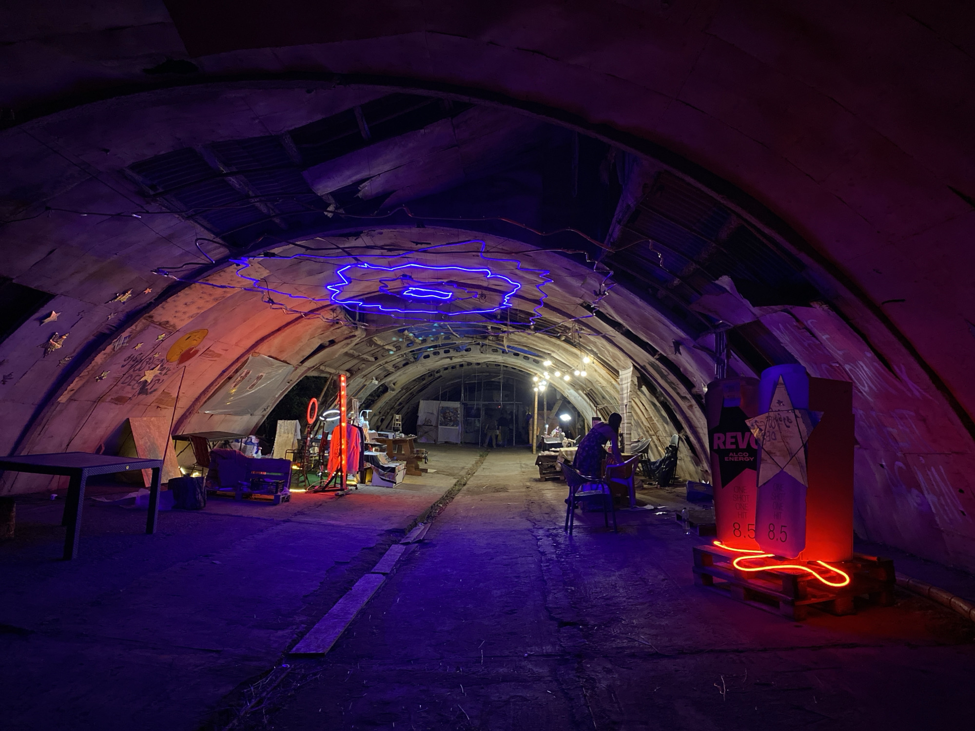 The image shows an interior space within an abandoned structure in Asureti, Georgia. The space is cavernous with a curved ceiling made of corrugated metal and concrete. The walls are covered in graffiti and art. The scene is illuminated by a combination of artificial lights, including blue and red neon signs.
In the foreground, a black table is positioned to the left. The floor is dirt or concrete. Further into the space, several individuals are engaged in various activities. To the left, there's a stand with red lighting and some clothing displayed. In the center and right, tables and chairs are set up, with people working or socializing. One person, a woman in a dark dress, is standing by a table on the right.
Dominating the right side of the frame is a large, cutout sculpture resembling a bottle or packaging, adorned with red neon lights that spell out "8.5". This sculpture is placed on wooden pallets. Overhead, strings of bare light bulbs provide general illumination. The overall atmosphere is one of an artistic or creative gathering taking place in a raw, industrial setting.