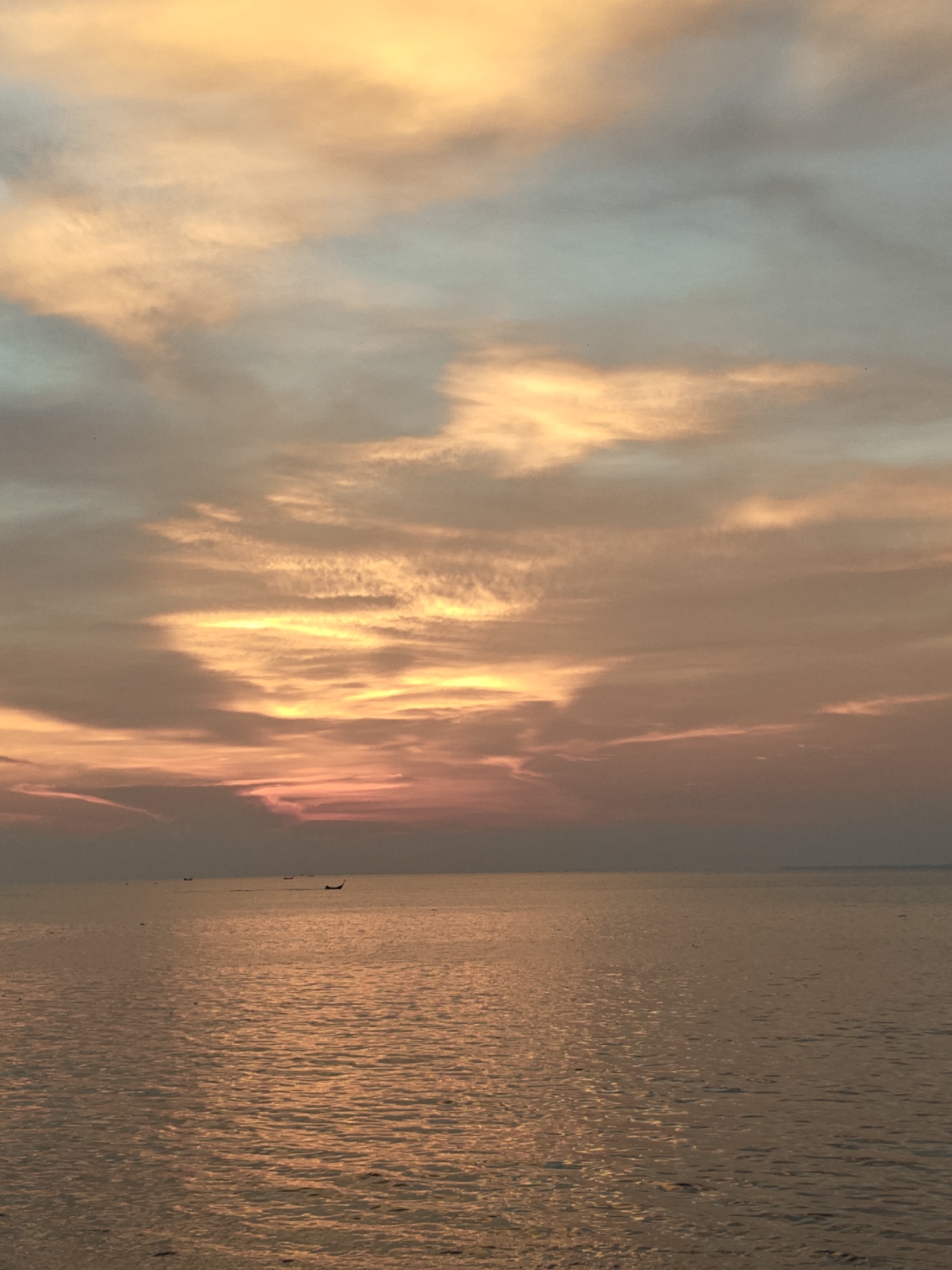 The image depicts a sunset over the ocean, presumably in Lhokseumawe, Indonesia, given the prompt. The sky is filled with clouds, predominantly in shades of orange, pink, and yellow, with some areas of blue. The sun appears to be setting near the horizon, casting a warm light across the scene. The ocean reflects the colors of the sunset, creating a shimmering effect. Several small boats can be seen in the distance on the water.