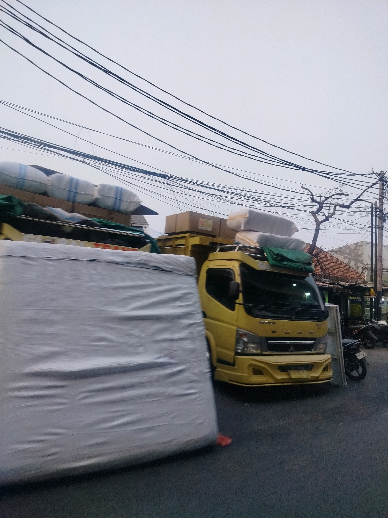 A yellow FUSO truck, laden with several large white cylindrical objects and boxes, is parked on a street. The truck's front is visible, featuring a prominent bumper and grille. Partially obscuring the truck from the left is a large, light-colored object wrapped in plastic sheeting, suggesting it's either cargo or part of another vehicle. The street is paved, and on the right side of the image, several motorcycles are parked. Buildings with tiled roofs and signage are visible in the background, indicating an urban environment. Overhead, a complex network of power lines crisscrosses the sky, which is overcast. The scene suggests a busy commercial or residential area in East Jakarta, Indonesia, likely during daytime.