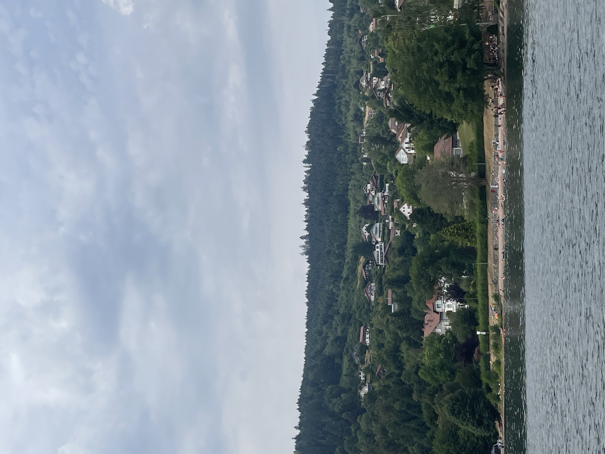 The image shows a lakeside scene in Gérardmer, France. In the foreground, a body of water with rippling surface is visible. A shoreline with a sandy beach stretches across the middle ground. Numerous people are present on the beach, some are sunbathing, while others are in the water, possibly swimming. Several orange buoys are floating on the water. Behind the beach, a densely forested hillside rises, dotted with residential buildings. These houses appear to be of various sizes and architectural styles, with visible roofs and windows. The hill is covered in lush green trees, forming a dense canopy. The sky above is overcast with a layer of gray clouds.