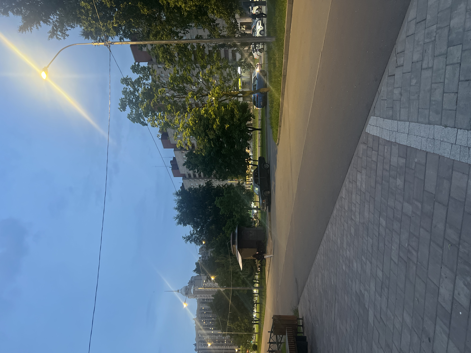 This image captures an urban street scene in Saint Petersburg, Russia, at dusk or early night, characterized by a deep blue sky with visible clouds and active artificial lighting. The location is identifiable by the distinctive dome and spire of the Petrovsky Passage (Knyaz Vladimir residential complex) visible in the distant left background, situating the scene likely in the Primorsky District, possibly near Kolomyazhsky Prospekt.
In the foreground, a paved sidewalk composed of gray rectangular bricks occupies the left, transitioning to an asphalt road on the right, delineated by a partial white block marking. A dark wooden bench is visible on the far left.
The midground features a wider asphalt road with a central grassy median strip populated by green-leafed trees. A single individual, dressed in dark pants and a lighter top, walks from left to right near a small, dark, kiosk-like structure with a sloped roof. Multiple vehicles are present on the road: a dark sedan is parked on the left, while further down the street, a blue SUV, a white car, and other light-colored vehicles are either parked or in motion, with some headlights or taillights visible. A prominent streetlamp with a bright, radiating yellow light stands on the right side of the frame.
The background consists of several multi-story residential apartment buildings with numerous illuminated windows, indicating occupancy. The architecture suggests post-Soviet era construction. Overhead, a thin power line stretches across the upper portion of the sky. The overall atmosphere is that of a quiet residential area transitioning from day to night.