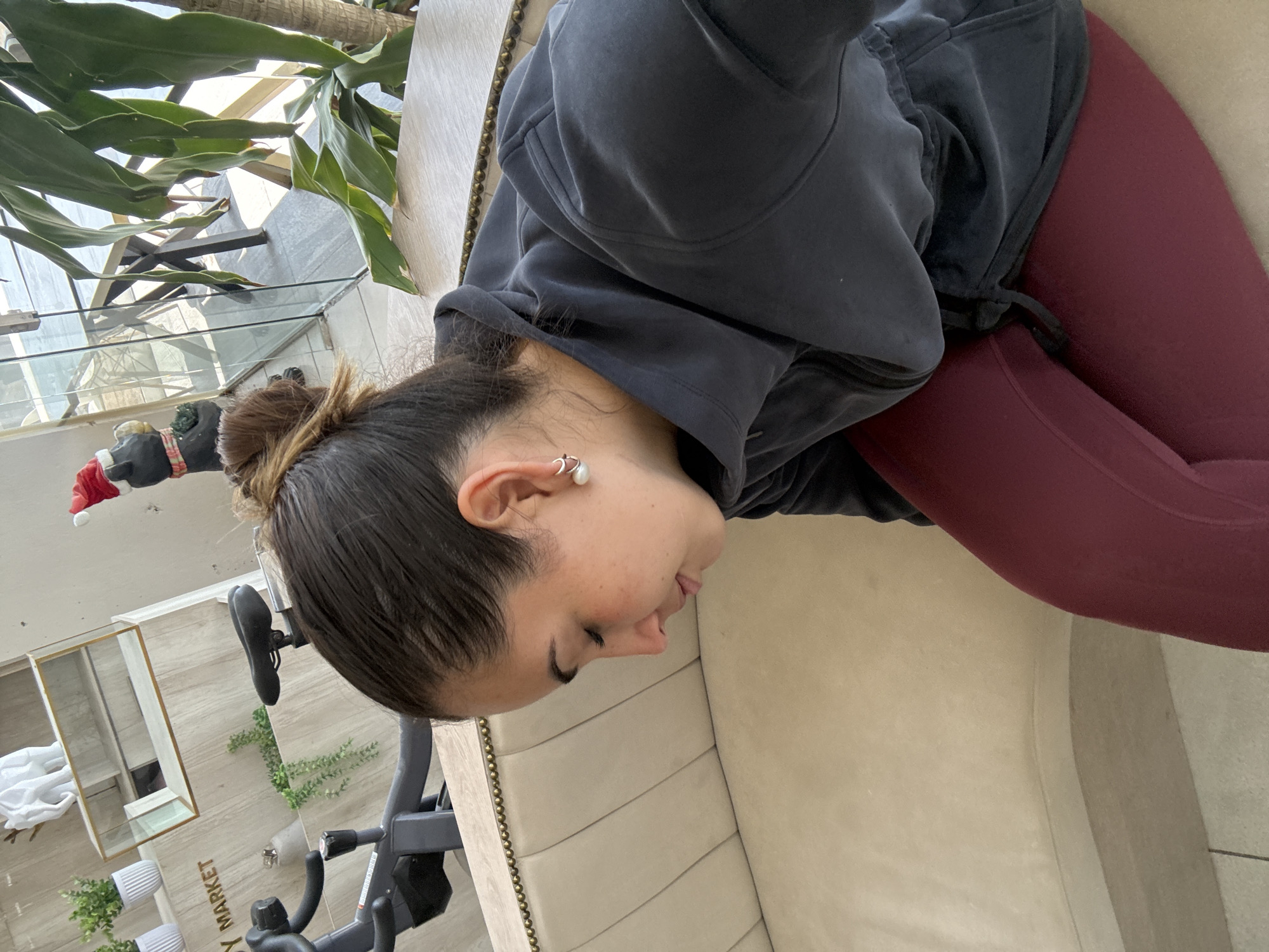 A female individual is depicted in a seated position, observed from the upper torso, facing downward and slightly to her right. Her dark hair is styled in a bun, and she exhibits visible freckles on her cheek. She wears a dark gray hooded jacket with a zipper closure and maroon-colored leggings. A silver hoop earring with a white, spherical dangle, possibly a pearl, is visible in her left ear.
She is seated on a light beige upholstered sofa featuring horizontal tufting and a line of brass stud detailing along its upper edge.
The background to the left reveals a black stationary exercise bike. Behind the bike, a light-colored wooden wall or paneling displays shelving. On these shelves are three small potted green plants in white cylindrical pots, a white decorative elephant statue, and a rectangular glass box with a gold-colored frame. A partial sign reading "Y MARKET" is visible on the wooden paneling below the shelves.
To the right of the subject, a large potted plant with broad green leaves is present. Beyond this plant, a clear glass partition, likely a window or balcony barrier, provides a view of a sunlit outdoor area with indistinct structures. A small black decorative figure, possibly an animal, is situated near the glass, wearing a red and white Santa hat and a red and white striped scarf.
The scene takes place in an indoor commercial setting, characterized by natural light. The "Y MARKET" sign strongly suggests the location is part of a "YAMA Healthy Market" establishment in Puebla, Mexico, which typically includes cafe or health-related amenities.