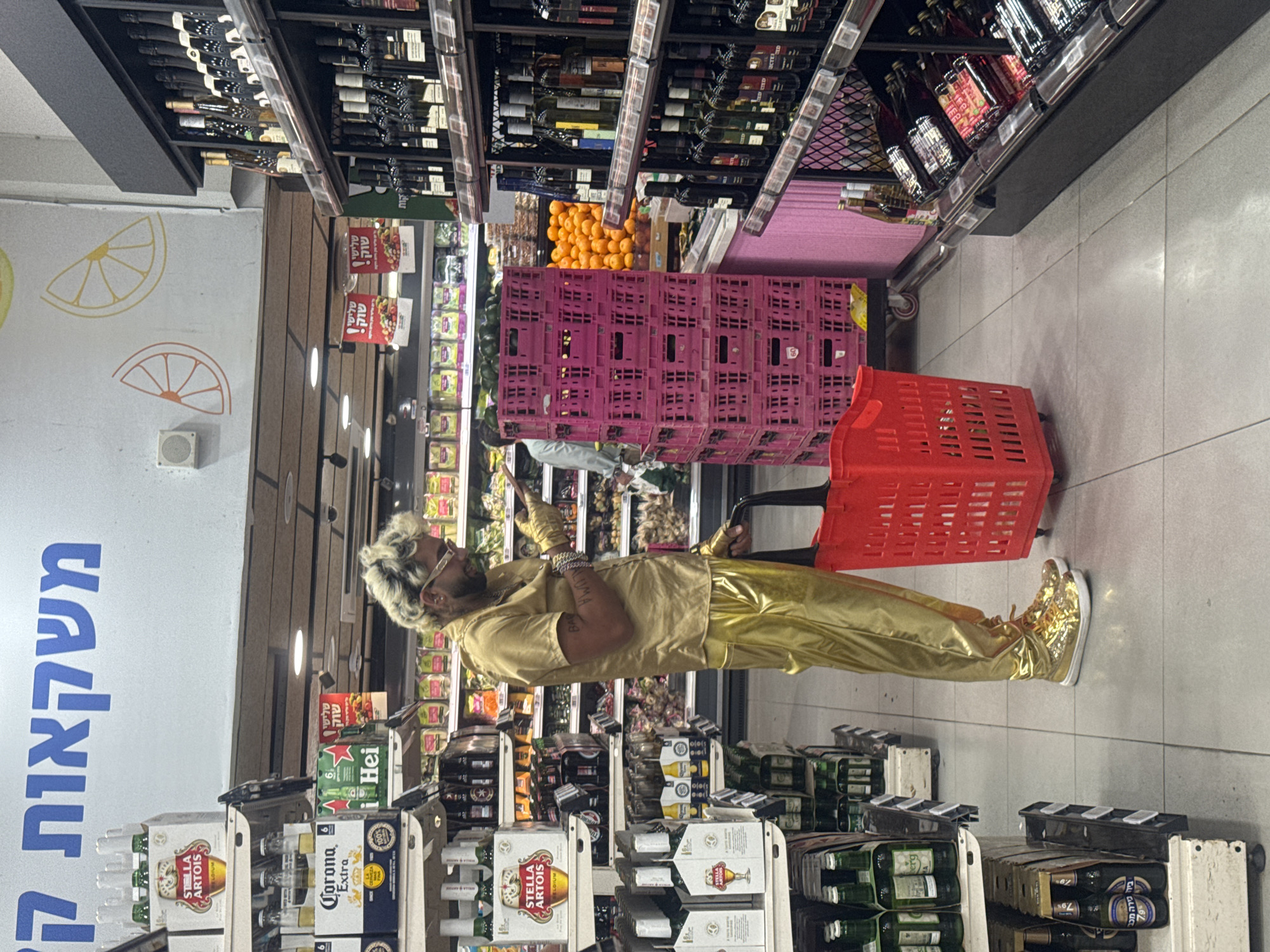The image captures a scene inside a supermarket. A person dressed in a shiny gold outfit, including shoes, pants, and a shirt, with blonde hair and sunglasses, is standing in an aisle with a red shopping cart. The person is facing towards the left, gesturing with their right hand. The aisles around the person are stocked with various products including alcoholic beverages like Stella Artois and Corona beer, as well as fresh produce like oranges. There are crates of pink-colored items stacked near the person. The store's wall features painted images of orange and lemon slices, and there is Hebrew text. The location is a supermarket in Givatayim, Israel.