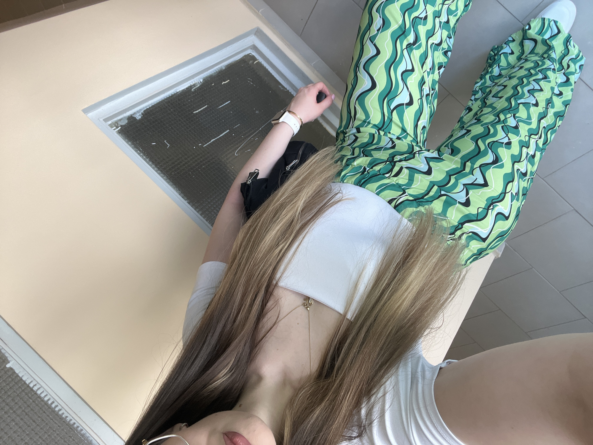 A close-up, angled shot shows a person lying on a beige surface, possibly a bench or low wall, against a light-colored wall with a square, recessed light fixture. The person is wearing green patterned pants with white shoes. They also have on a white top, a wristwatch, and a thin necklace with a pendant. A black bag rests next to their head. Their long, brown hair is partially covering their face. The exact location in the city of Unknown, Unknown is unidentifiable.