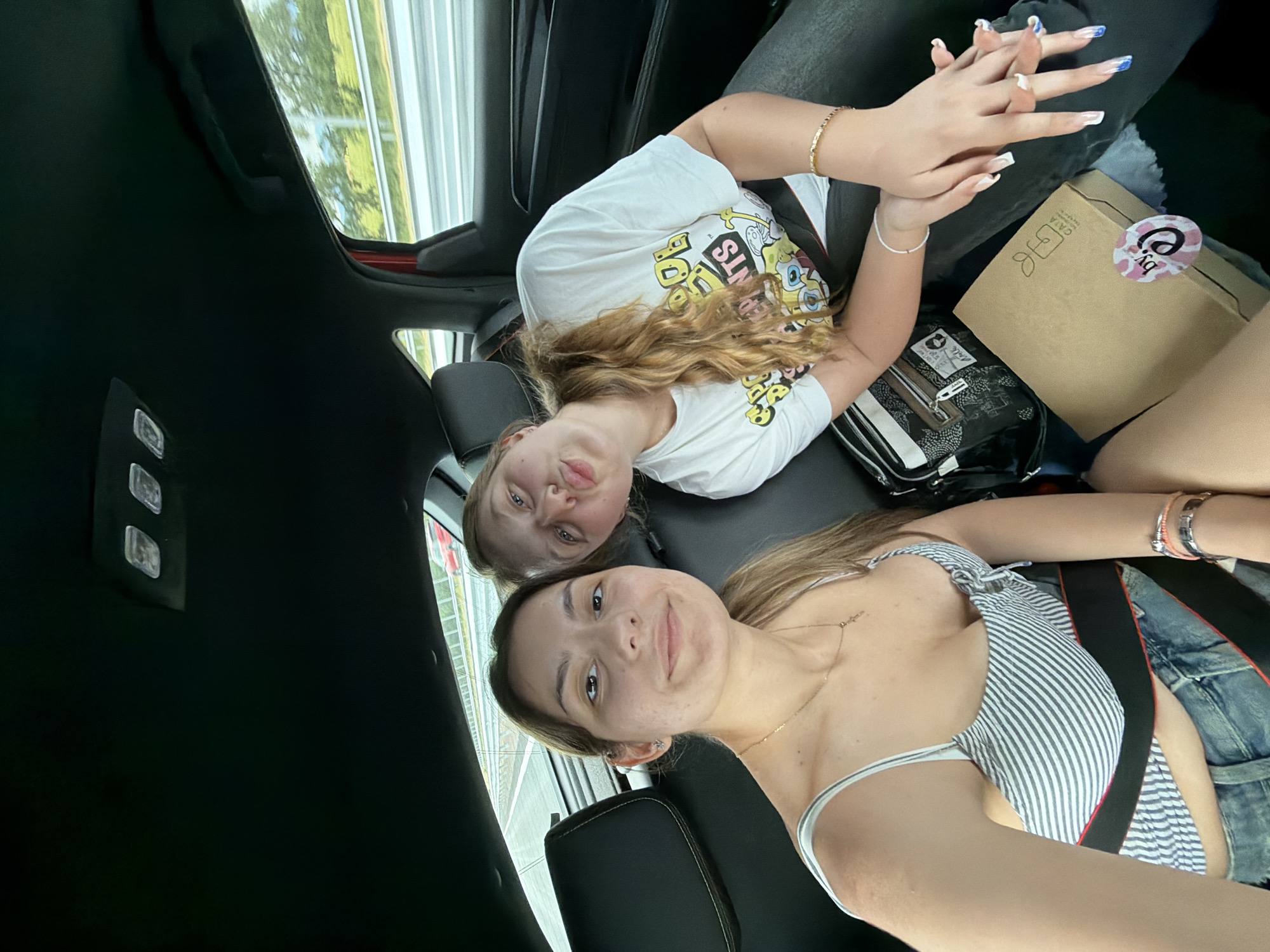 Two young women are inside a vehicle, possibly a car. The first woman, closest to the camera, is wearing a striped top and jeans with a visible seatbelt. She has light brown hair and a necklace. The second woman, behind her, is wearing a white t-shirt with a graphic design. She has blonde hair. Their hands are clasped together. Several items are on the seat, including a black and white patterned bag and a brown box with a sticker. The background shows a blurred view of trees and structures outside the car window. There is no indication that the image was taken in Chokoba, Bulgaria.