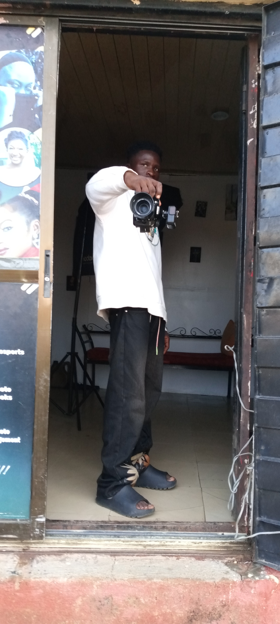 A young person, likely male, with dark skin and short, dark hair, stands in a doorway, holding a black camera with a prominent lens and an attached external monitor directly towards the viewer. He is dressed in a white long-sleeved top, black trousers with a visible colorful drawstring, and dark open-toed slide sandals, revealing his bare feet.
The doorway leads into an interior space with white walls and light-colored tiled flooring. Inside, a dark metal bench with a red cushion is visible against the far wall, alongside a folded metal stand and some small framed pictures. The ground immediately outside the doorway is a reddish-brown color, suggesting dirt or worn concrete.
To the left of the doorway, a dark sign features several portrait images of women and partial text. Visible fragments of text include "...sports", "...oto", "...ooks", "...oto", and "...ajement", indicating the establishment might be a photography studio or offers related services like passports, photo books, or photo management. White electrical cords hang from the right side of the door frame. The lighting suggests it is daytime. The scene depicts a commercial setting, possibly a photography studio, in Jos, Nigeria.
