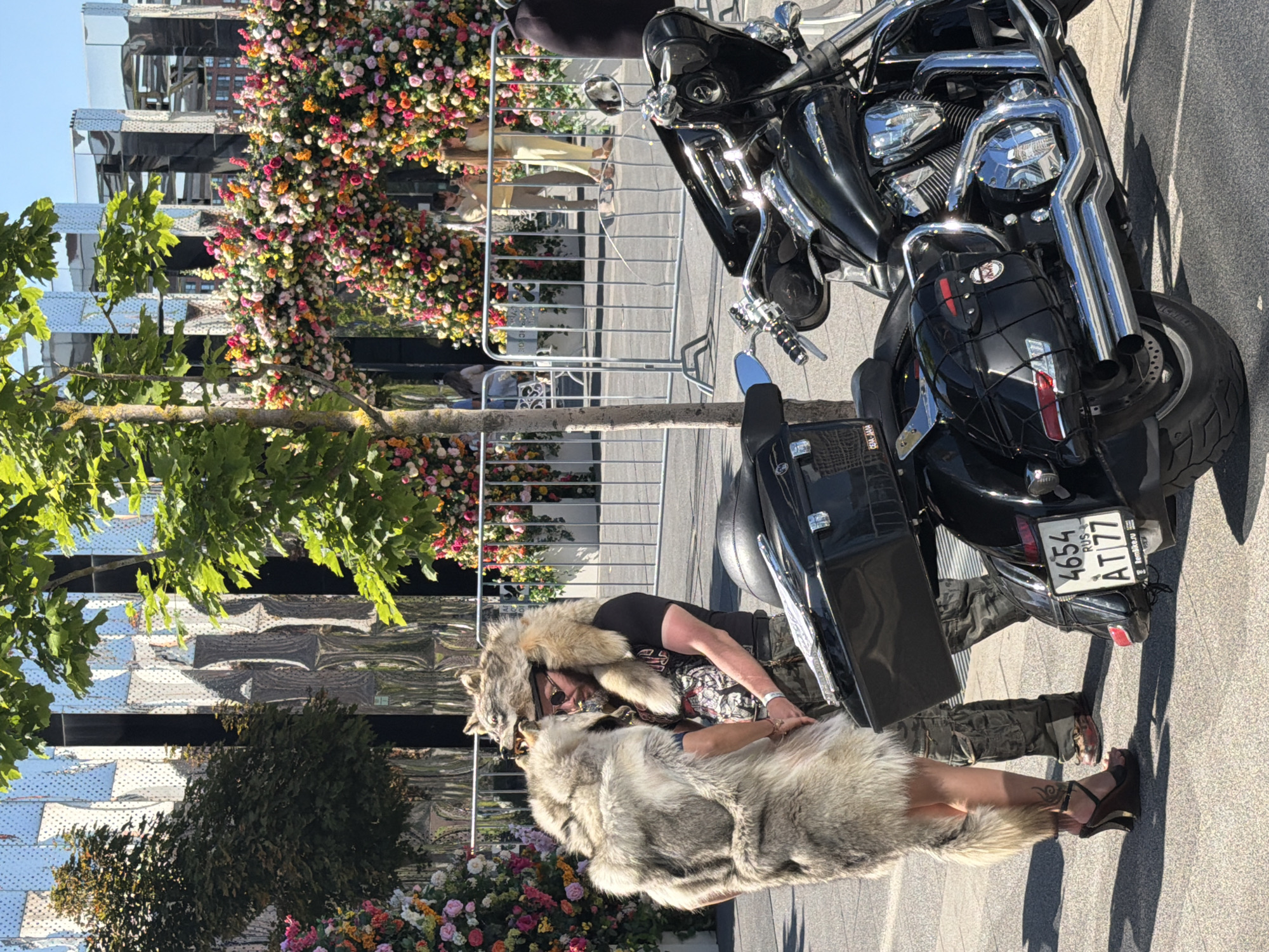 The image depicts an outdoor urban scene in the city of Khimki, Russia, under bright daylight. In the foreground, a black cruiser-style motorcycle is parked on a paved surface. Its visible components include chrome exhaust pipes, engine casing, a black seat, saddlebags (one with a cargo net), and a windshield. The rear license plate reads "4654 AT 77 RUS" with "RemBike.ru" on the frame.
Two individuals stand near the motorcycle. The person on the left, viewed primarily from the back, appears to be wearing a light-colored fur garment resembling an animal pelt, covering their upper body and extending to the mid-thigh, with a prominent tail-like section. This individual wears dark, high-heeled wedge sandals, revealing a tattoo on their right ankle. The person on the right is a man with a beard and sunglasses, wearing a black t-shirt with a graphic design and camouflage cargo pants. He is wearing a fur hat or garment fashioned from a wolf's pelt, with the wolf's head positioned over his own head and the fur draped over his shoulders. Both individuals are positioned facing each other, suggesting an interaction.
Behind the individuals and the motorcycle, silver-colored metal crowd control barriers are present. Beyond these barriers, large, multi-colored floral arches, densely covered with various flowers in shades of pink, orange, yellow, and white, form a decorative display. In the background, a modern building features a highly reflective, patterned facade, suggesting a metallic or mirrored surface. Green-leafed trees are visible on the left and right sides of the frame. The ground is paved with light-colored interlocking pavers, and distinct shadows indicate a sunny environment.