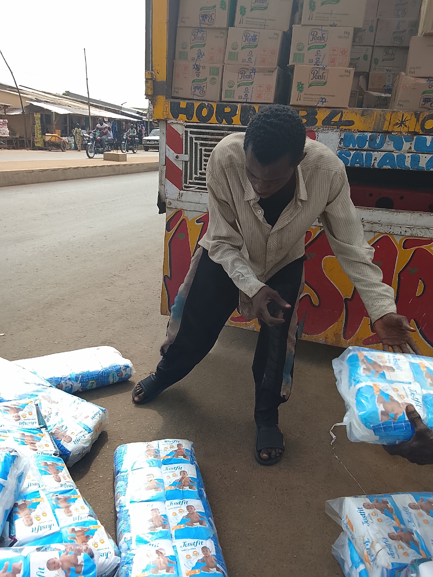 In Akwanga, Nigeria, a Black man in a light brown striped shirt and dark pants is crouched down next to a truck, holding a package of "Justfit Baby Diapers." A pile of similar diaper packages is on the ground in front of him. The truck is partially visible, with boxes of "Peak Full Cream Instant Milk Powder" stacked inside and colorful graffiti on its side. In the background, market stalls with corrugated metal roofs line a dusty street where two men on motorcycles are riding past. A white car is also visible on the road. The scene appears to be a street-level commercial area in the city.