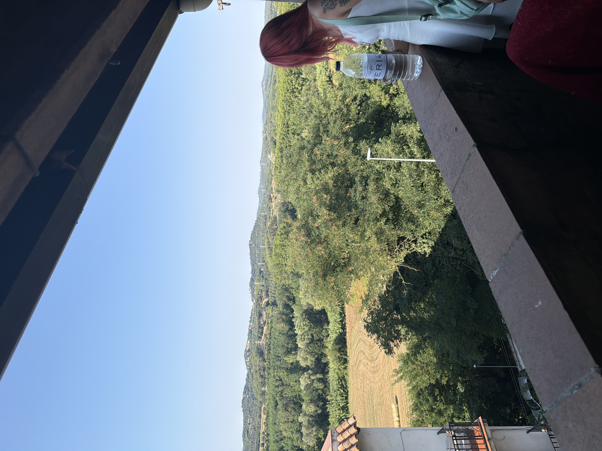 A woman with vibrant red hair stands on a balcony overlooking a lush green landscape in Hostalric, Spain. She is wearing a white dress and a light green bag is slung over her shoulder. In her right hand, she holds a clear plastic water bottle with a label that reads "ERIA". The balcony railing is made of brick. In the background, rolling hills are covered with trees, and a field with harvested crops and hay bales is visible in the mid-ground. The sky is clear and blue. A portion of a building with a red tile roof is on the left side of the frame.