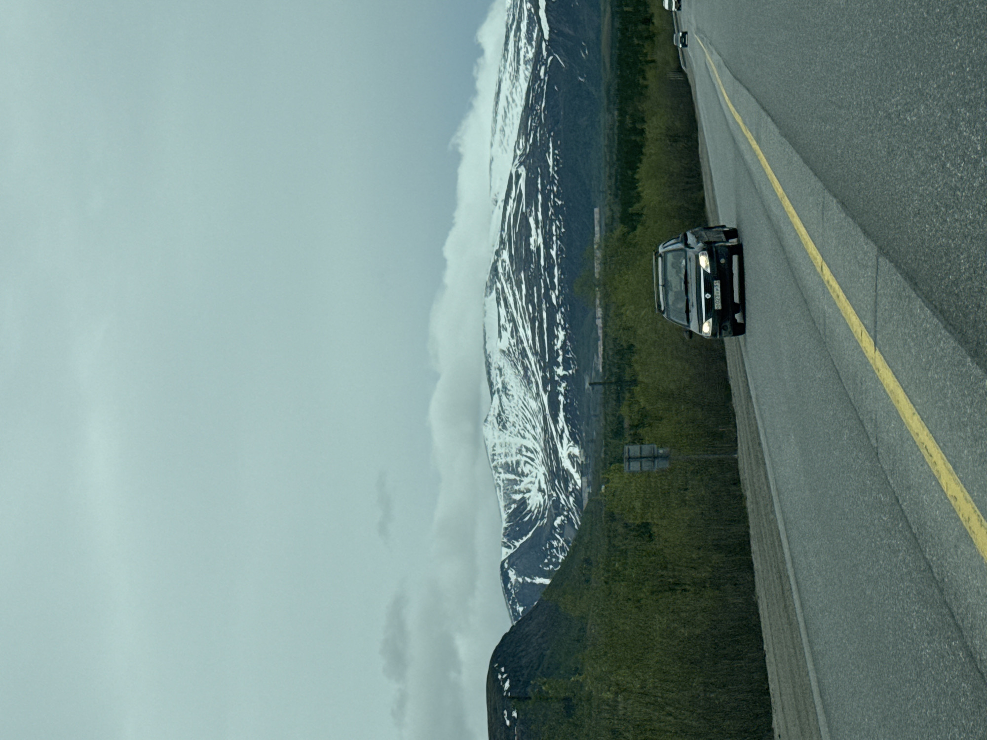 The image shows a road next to a mountain range. A black car is traveling on the right lane of the road. A yellow line divides the lanes. There is a sign on the side of the road. The sky is overcast. The mountain range is partially covered in snow. There is vegetation on the lower slopes of the mountain. There is no indication that this image is located in Apatity, Russia.
