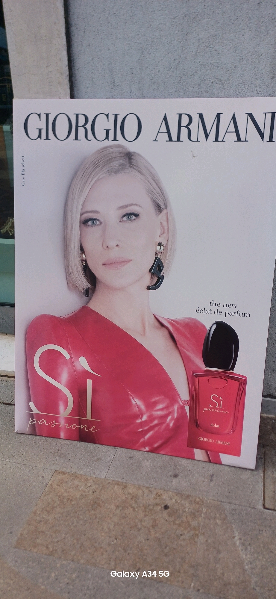 A full-frame photograph shows a large advertisement poster for Giorgio Armani's "Si Passione" perfume, prominently featuring actress Cate Blanchett. The poster is positioned outdoors, leaning against a light-colored textured wall that appears to be made of concrete blocks, with a paved ground surface in the foreground. The ground is composed of large, irregularly shaped stone slabs with a light brown hue, typical of pedestrian walkways.
Cate Blanchett, with her signature blonde bob and blue eyes, is depicted looking directly at the camera. She is wearing a vibrant red, shiny garment that resembles leather or patent leather. Her makeup is subtle, with emphasis on her eyes and lips. She is adorned with a striking pair of black, abstract-shaped earrings.
To the right of the image, a bottle of "Si Passione" perfume is displayed. The bottle is rectangular with rounded edges and has a glossy red finish, echoing the color of Blanchett's attire. The label on the bottle features "Si passione" in gold lettering, with "éclat" and "GIORGIO ARMANI" printed below. Above the perfume bottle, text reads "the new éclat de parfum."
The poster itself is white with large, dark sans-serif font for the "GIORGIO ARMANI" branding at the top. On the left side of the poster, vertically oriented text reads "Cate Blanchett."
The lighting suggests it is daytime, with no harsh shadows, indicating possibly an overcast sky or indirect sunlight. The overall scene is a street-level view of an advertisement in a public space. The photograph was taken with a Samsung Galaxy A34 5G phone, as indicated by the watermark in the bottom right corner. The location is Abano Terme, Italy, as stated in the prompt.