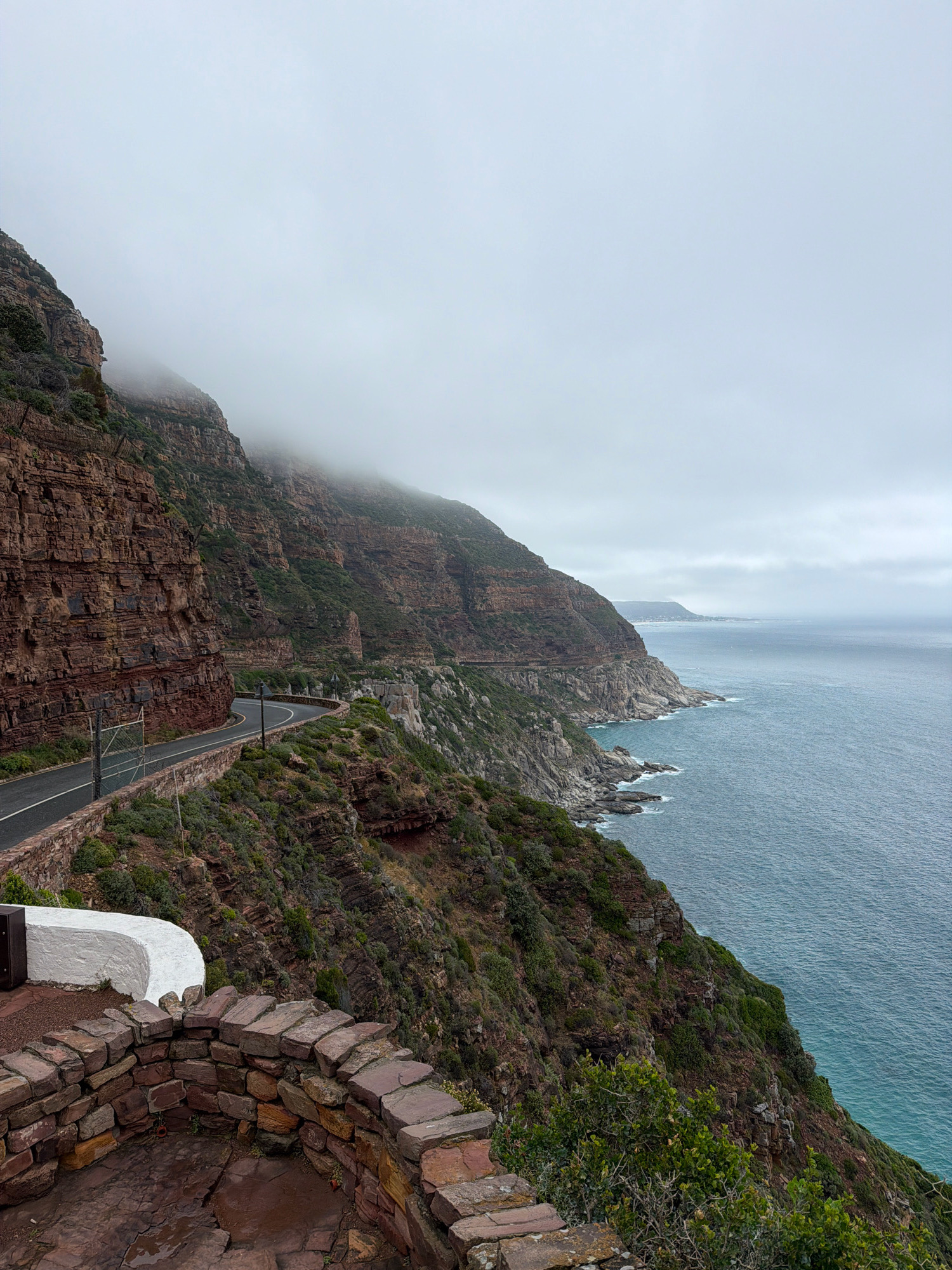 The image depicts a scenic coastal road carved into a steep, rocky cliff face overlooking a vast expanse of the ocean. The road, with a single lane and white markings, curves sharply along the cliff's edge. A low stone wall with a white concrete cap runs parallel to the road, separating it from the precipitous drop. The cliffs are characterized by layered, reddish-brown rock formations interspersed with patches of green vegetation clinging to the slopes.
In the foreground, a circular stone-walled viewing area with a reddish-brown paved floor is visible. The stones are roughly hewn and stacked, creating a rustic barrier. Beyond this area, the dramatic landscape unfolds. The ocean is a deep blue-green, with gentle waves lapping against the rocky shoreline in the distance. The sky is overcast and hazy, with a band of thick fog obscuring the upper portions of the cliffs and the horizon.
In the far distance, a coastal town or settlement can be faintly seen nestled along the coastline. The overall atmosphere is one of rugged natural beauty and a sense of isolation, amplified by the overcast weather. This vista is characteristic of the Chapman's Peak Drive, a renowned scenic route located in Hout Bay, South Africa, known for its breathtaking cliffside views. There are no people or vehicles visible in the frame.