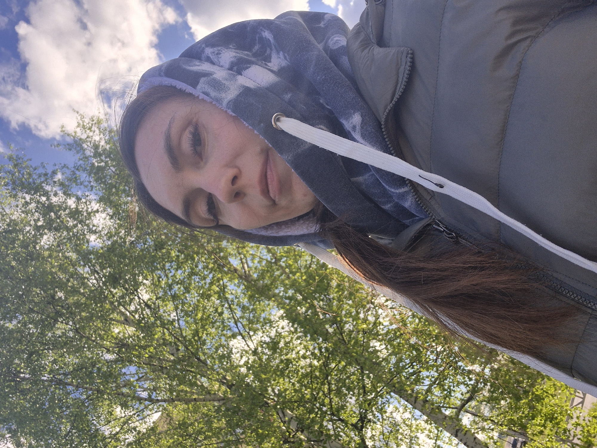 A selfie was taken from a low angle by a woman with long brown hair, wearing a gray hooded jacket, outdoors during the day. The background shows trees with light green leaves under a bright sky with scattered clouds. This image was taken in the city of Chaikovsky, Russia.
