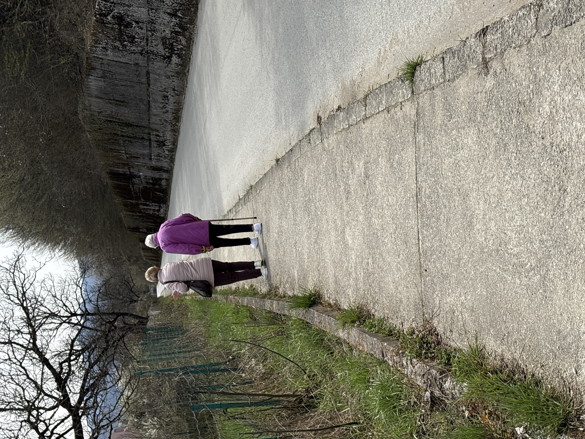 Two elderly women are walking on a paved path next to a concrete wall on the outskirts of Teteven, Bulgaria. The woman in front wears a beige jacket and carries a dark purse over her shoulder. The woman behind wears a purple jacket and uses a walking stick. The path is bordered by vegetation and trees. A tall concrete structure stands beside the path.