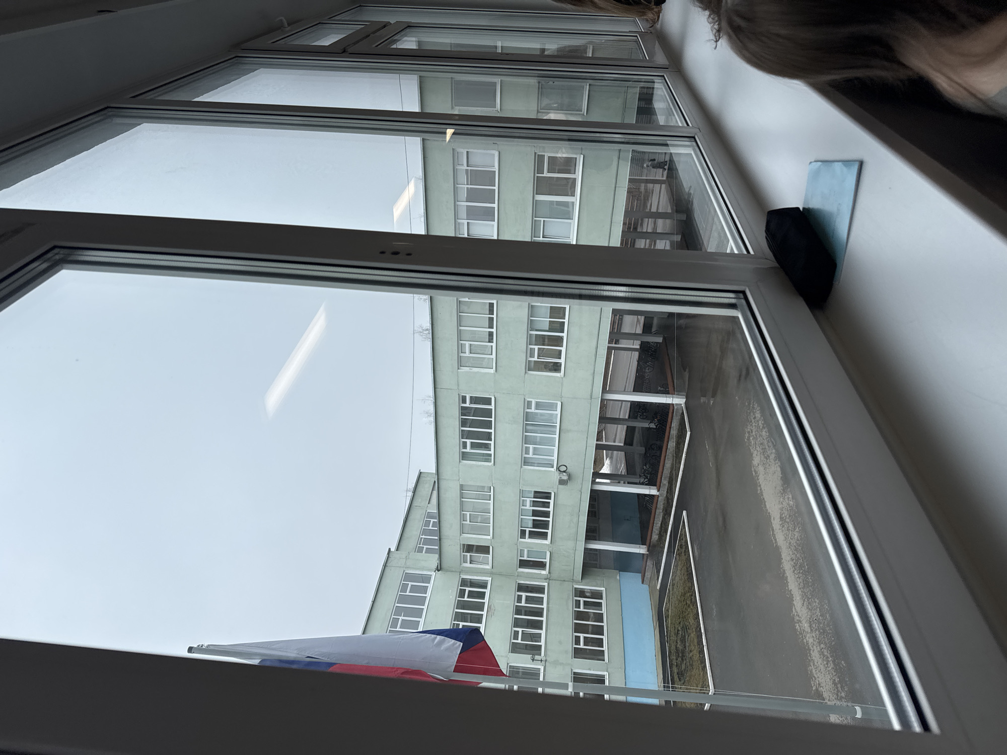 A view from a window of a building in Chaikovsky, Russia, looking towards another multi-story building with light green facade. The building outside has many windows and a covered walkway. A Russian flag is partially visible at the bottom left. A black object and a blue sticky note are on the window sill. The sky is overcast.