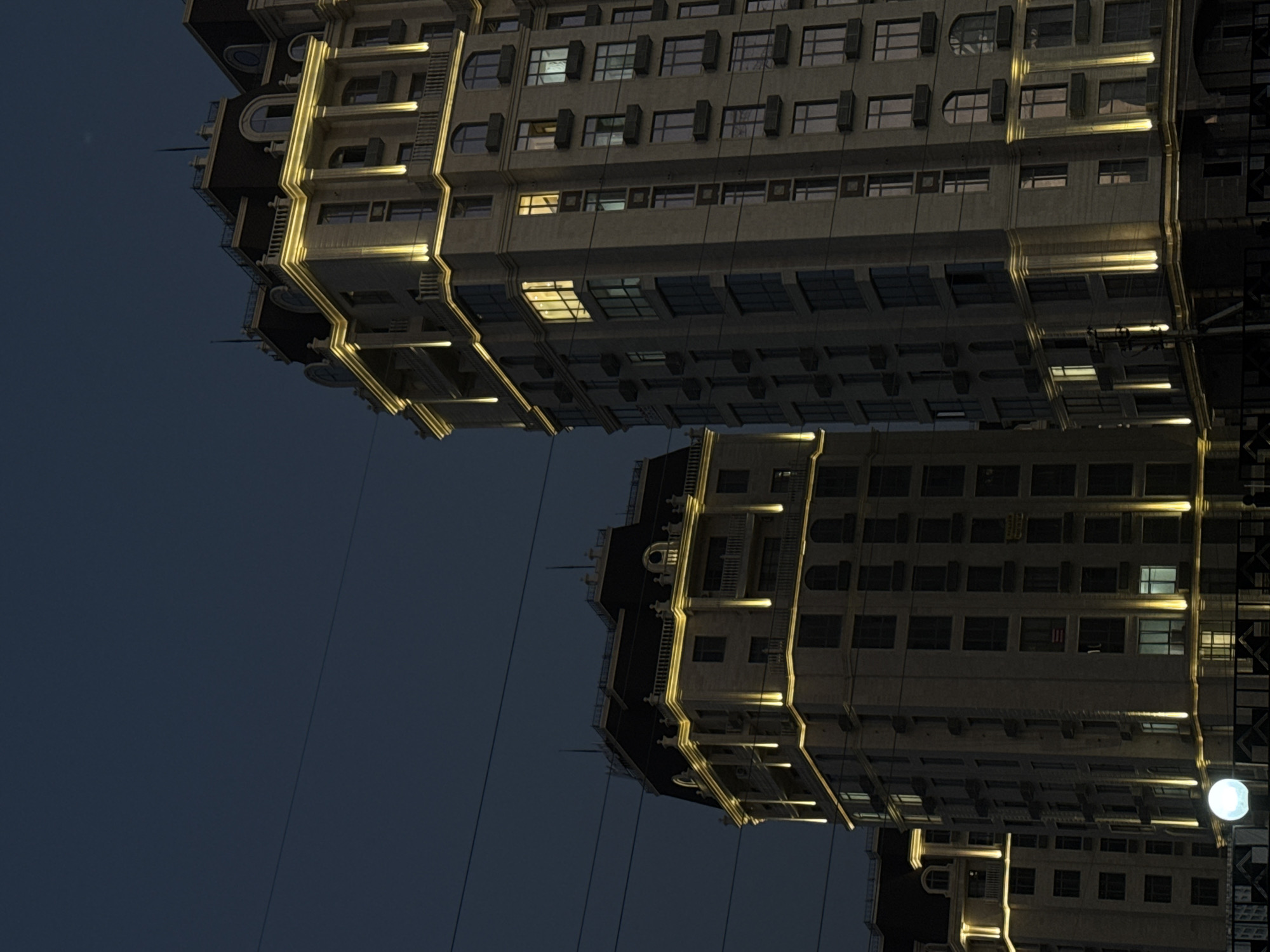 Two tall, modern apartment buildings with ornate architectural details stand illuminated against a dark night sky. The buildings are primarily light-colored with dark mansard-style roofs topped with decorative railings. Yellow lights highlight the cornices and vertical architectural elements of both structures. The building on the right is taller and closer, revealing numerous windows, some of which are lit from within, suggesting occupancy. Several windows on the taller building have black planters. Power lines are visible crisscrossing the sky in front of the buildings. In the foreground, a bright, round light fixture is visible on the left, near the base of the buildings. The overall impression is of a contemporary urban setting at dusk or night. The architecture of the buildings is consistent with newer developments in Bishkek City, Kyrgyzstan, particularly in areas with modern residential complexes.