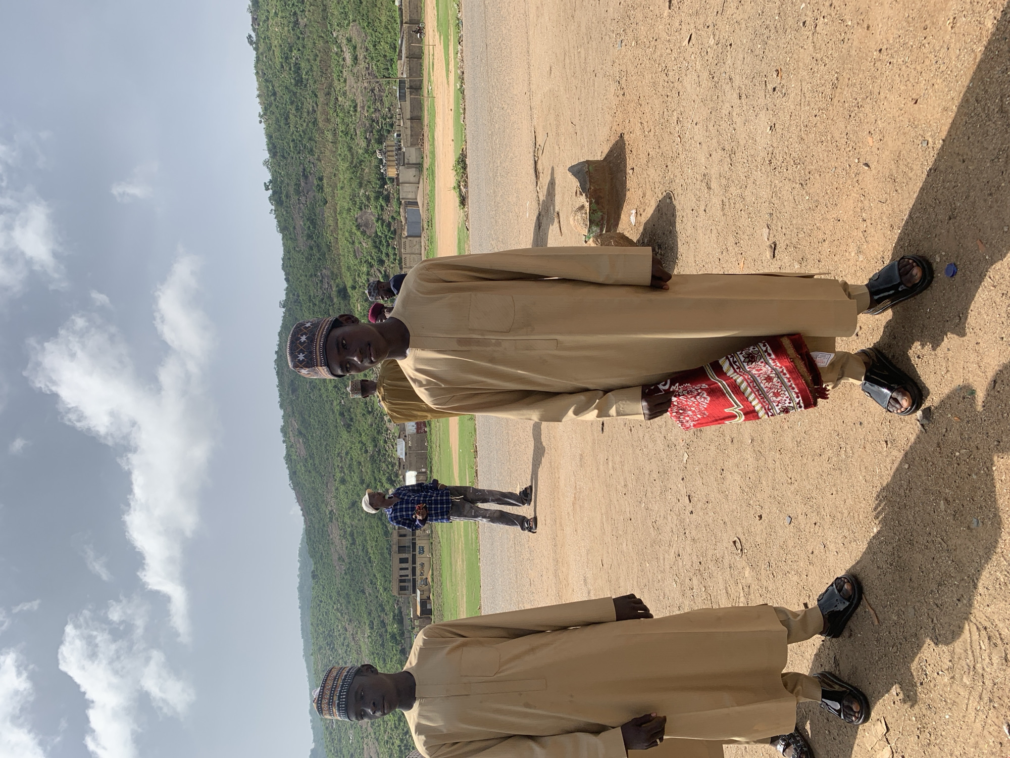 The image depicts several men in a public space in Naraguta Hausawa, Nigeria. The men are wearing traditional garments, including caps and long robes. One man holds a red prayer mat. They stand on a dirt ground. A building under construction is visible in the background. A green, hilly landscape is also present. The sky is partly cloudy.