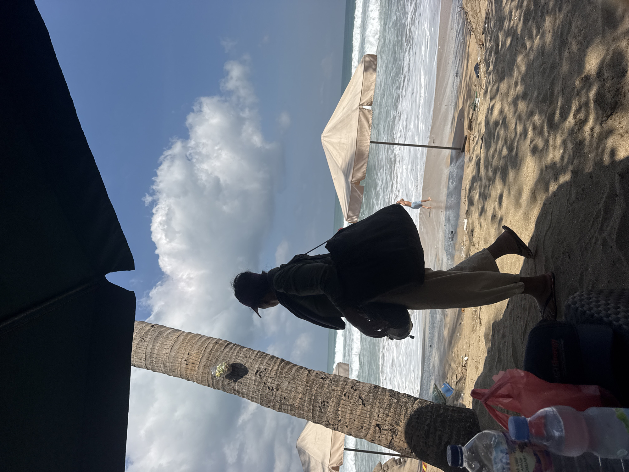 A person stands on a sandy beach in Kuta, Indonesia, looking out at the ocean. The person is wearing a dark jacket, a cap, and light-colored pants. They are carrying a large dark bag over their shoulder and a smaller bag in their hand. In the foreground, there are two plastic water bottles and a red plastic bag on a woven mat. A large palm tree trunk is on the left side of the image, and part of a black umbrella is visible at the top. In the background, there are two light-colored beach umbrellas and waves are breaking on the shore. A child can be seen playing in the shallow water near one of the umbrellas. The sky is blue with scattered white clouds.