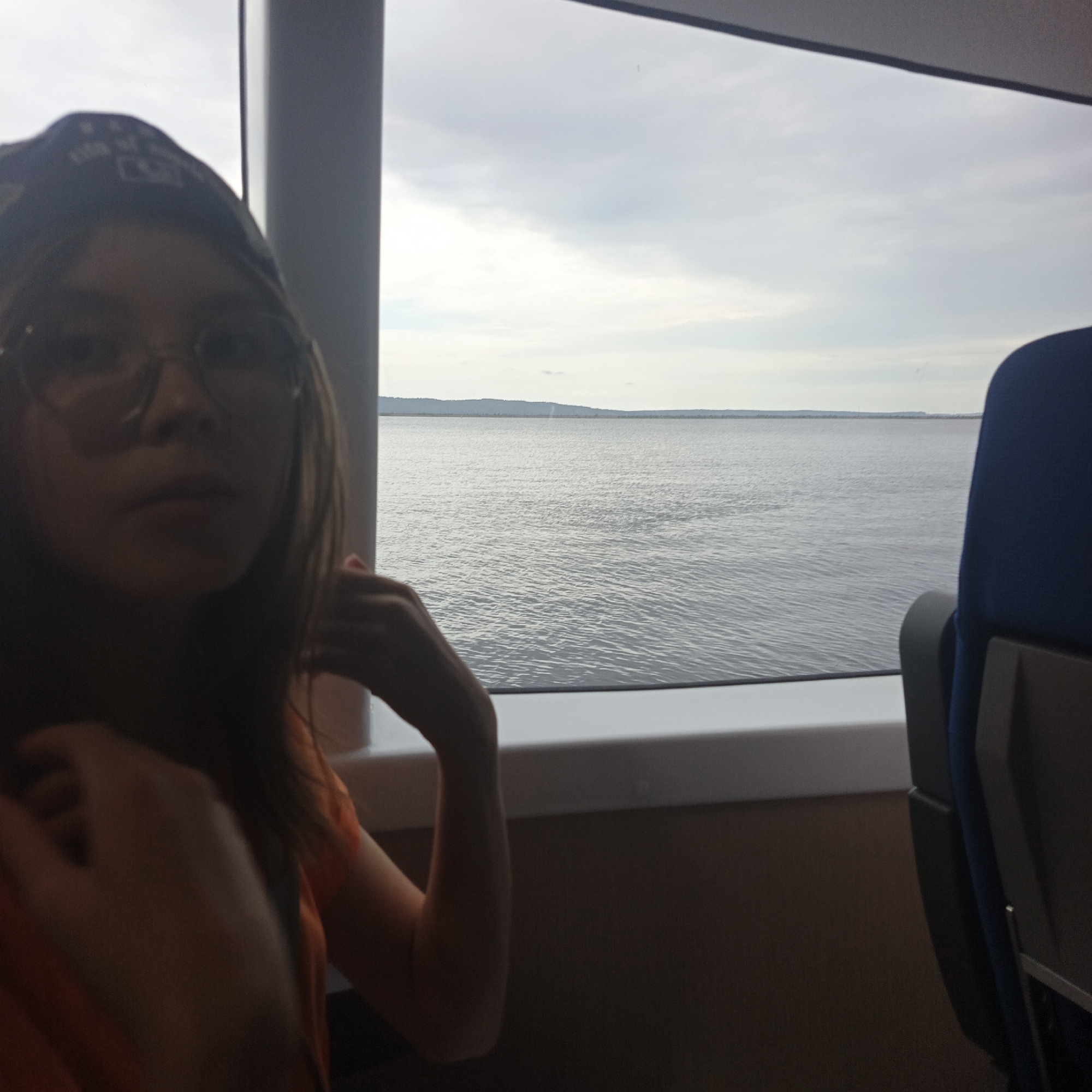 A young person wearing glasses and a baseball cap looks at the camera, partially obscuring their face. They are seated indoors, with a large window to their right offering a view of a calm body of water under a cloudy sky. The interior appears to be a mode of transport, possibly a train or ferry, as suggested by the blue seat visible on the right side of the frame. The person's left hand rests on their arm, and the right hand is positioned near their head. The overall lighting suggests an overcast day.