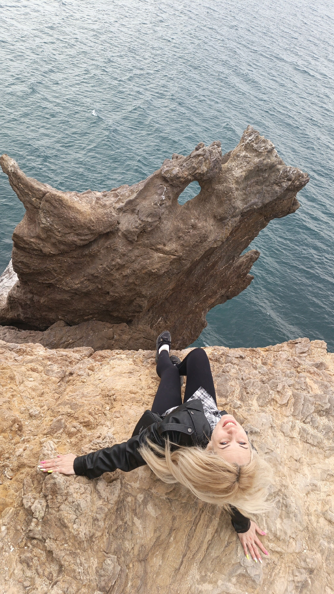 A young blonde woman lies on her back on a rocky cliff edge, looking up at the camera with a joyful smile. Her long, light-colored hair is fanned out around her head. She is dressed in a black leather jacket worn over a black and white plaid shirt, black leggings, and black shoes. Her arms are spread slightly, and her fingernails are painted in bright, multi-colored neon shades.
Below and to the right of the woman, a remarkable natural rock formation juts out into the dark blue-green, slightly choppy sea. This formation, resembling a mythical creature like a dragon's head with a prominent hole or archway through it, adds a dramatic element to the coastal landscape. The scene is set outdoors, likely during the daytime with diffuse lighting, suggesting an overcast sky. The woman's relaxed pose and happy expression convey a sense of adventure and enjoyment of the scenic location. The setting is a rugged, beautiful coastline in Балаклавский округ, Ukraine. No visible text is present in the image.