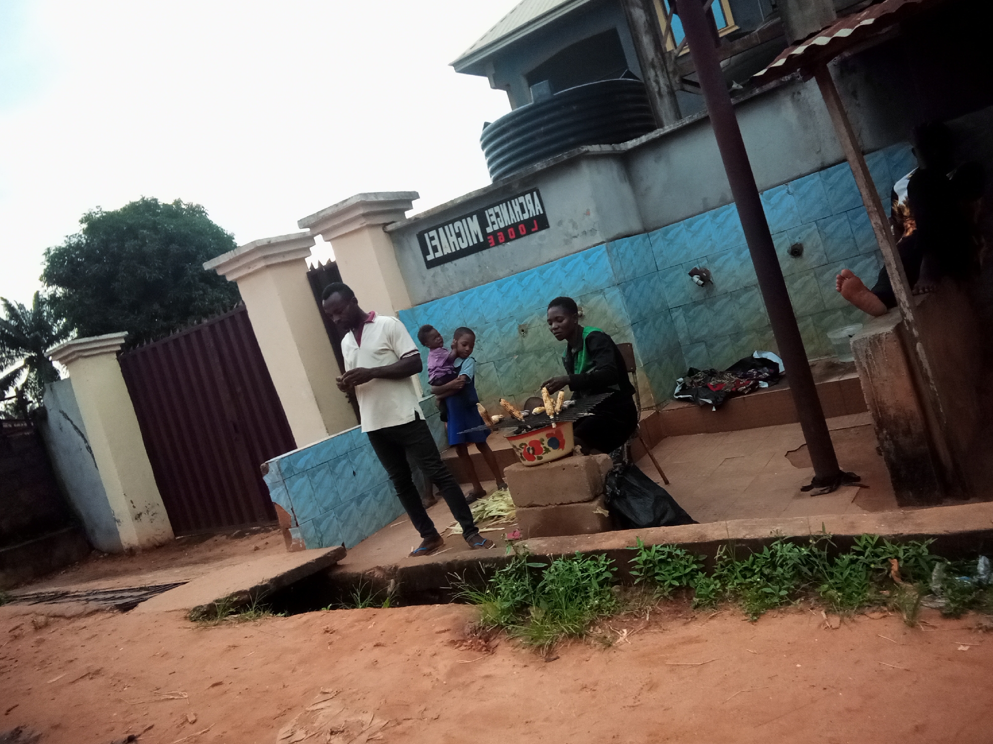 In Oko Town, Nigeria, this image depicts an outdoor scene where a group of people are gathered. A man in a white polo shirt and black pants stands to the left, facing away from the camera, holding a phone. Beside him, a woman in a green garment holds a child. Another child, dressed in a blue dress, stands between them. They are positioned near a barbecue grill where corn is being cooked. The grill is placed on a stack of concrete blocks, and a decorative bowl with a floral pattern sits beneath it. Scattered around the area are corn husks and a black plastic bag. In the background, a building with a large water tank is visible, along with a sign that reads "ARCHANGEL MICHEAL LODGE". The ground is covered in red dirt, with some patches of grass growing along a concrete edge. The overall atmosphere suggests a casual gathering or informal food preparation activity.