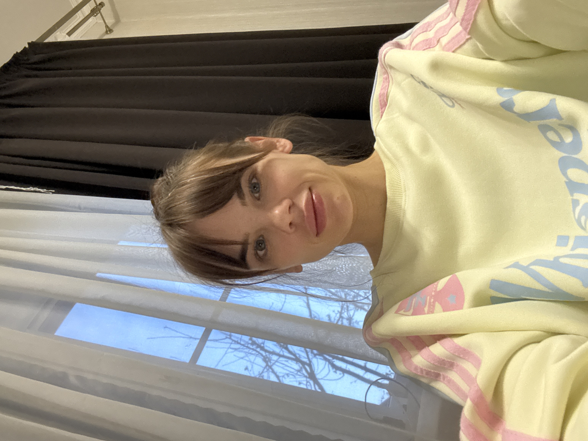 An indoor portrait-oriented image shows a young woman looking directly at the camera with a soft, pleasant smile. She has light brown hair styled with bangs and blue eyes. She is wearing a light yellow long-sleeved sweatshirt. The sweatshirt features light blue text, partially visible as "per" or "ee" on her chest, and light pink and white striped patterns on the shoulders and upper arms.
The background primarily consists of a window on the left side, covered by sheer, light-colored curtains through which a clear blue sky and the silhouettes of bare tree branches are visible. To the right of the woman, and partially behind her, are dark, pleated curtains, possibly black, which are drawn to the side. The lighting suggests it is daytime. The scene conveys a relaxed, casual atmosphere in what appears to be a residential setting. The location context is Rostov-on-Don, Russia.