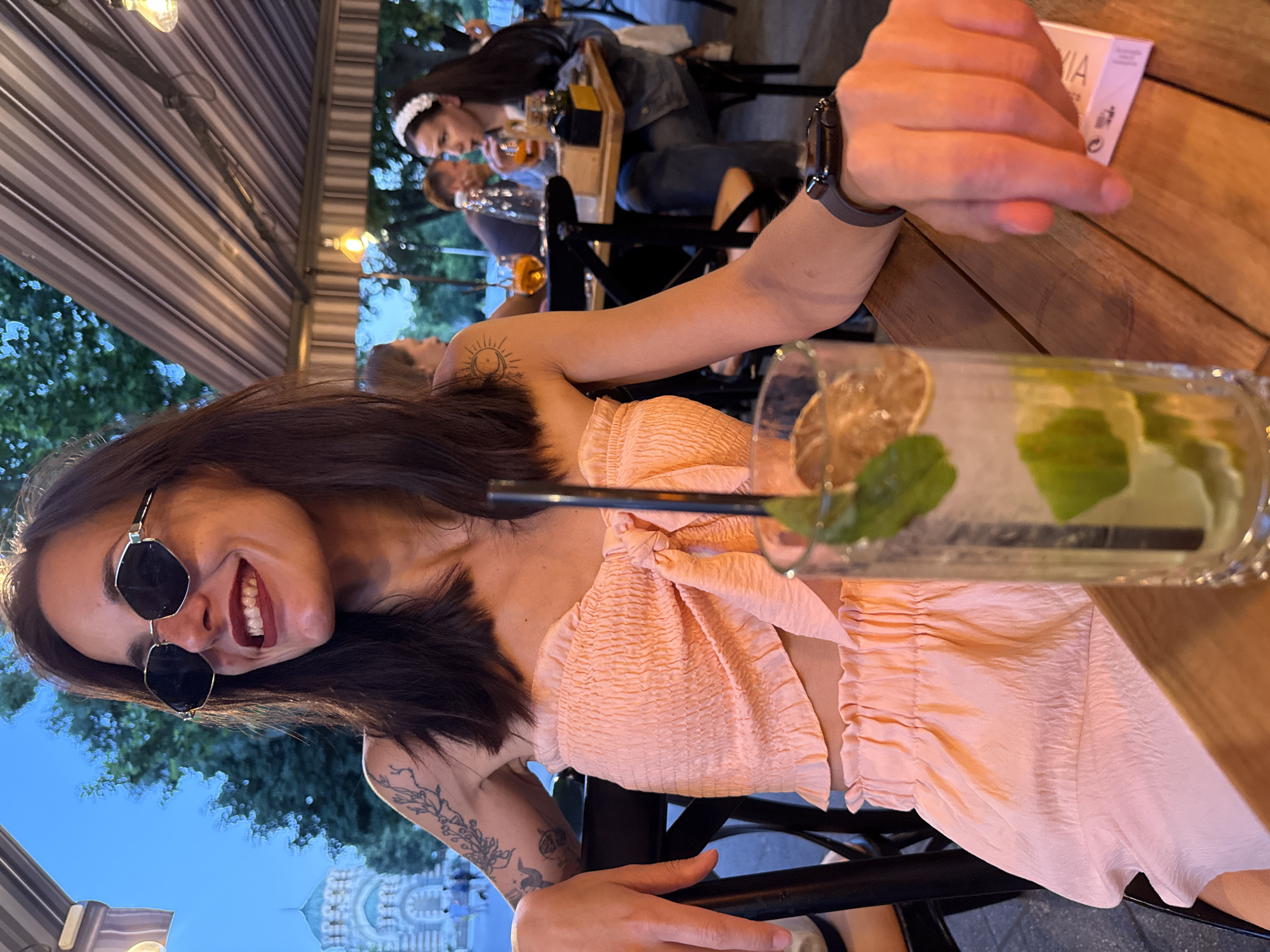 The photograph captures a woman seated at an outdoor table in Pleven, Bulgaria, likely at a restaurant or cafe. She is positioned in the foreground, facing the viewer's left, with a slight smile and her head turned away. She is wearing sunglasses with a hexagonal frame and a pale pink, textured bandeau top with a tie detail at the bust. On her left arm, there is a tattoo of botanical elements and celestial symbols. A tall glass containing a beverage with ice, lime, and mint is placed on the table in front of her, close to her right hand. Her right hand rests on the table, and she is wearing a dark-colored smartwatch on her wrist.
In the background, other patrons are visible at tables, some with drinks. The setting appears to be under an awning with striped fabric. To the far left of the frame, a building with a dome, resembling a church or historical monument, can be seen in the twilight. The overall atmosphere suggests a social gathering during the evening.