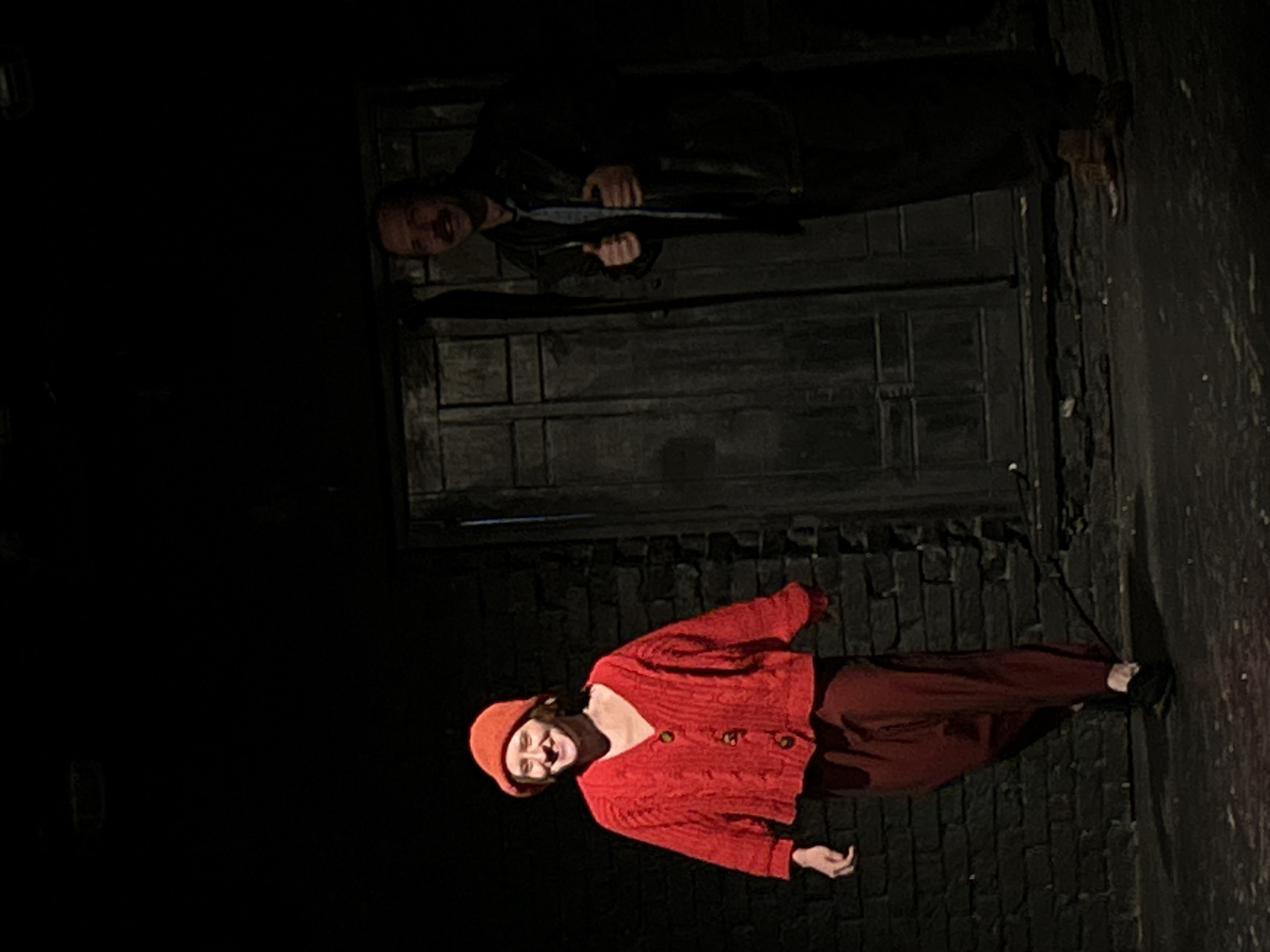 The image shows two people in front of what appears to be a brick wall and dark double doors. One person is wearing a red sweater, hat, and pants. The other person is wearing a dark jacket and pants. It's night, and the lighting is dim. The location is in Unknown, Unknown.