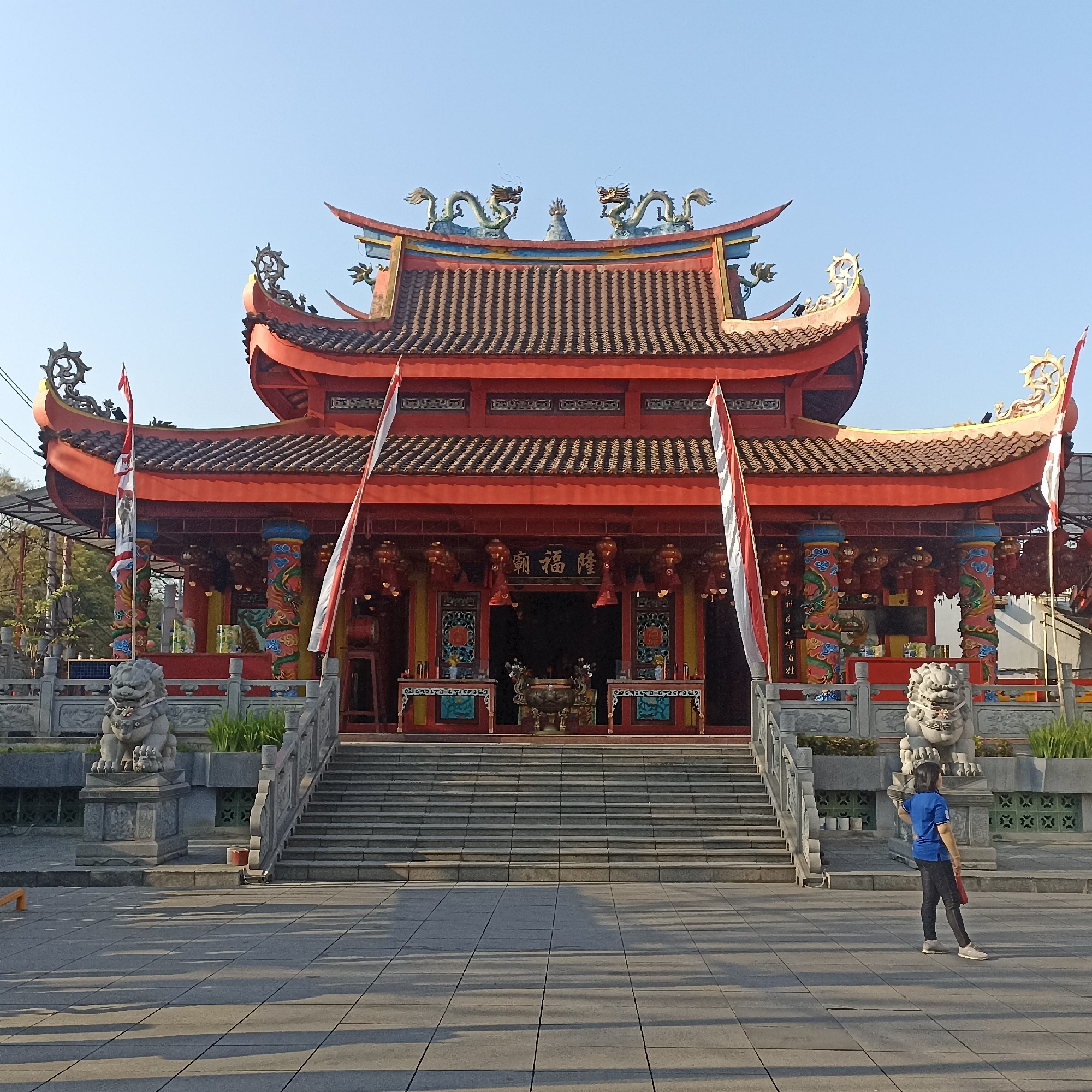 The image depicts the entrance of a Chinese temple located in the city of Magelang, Indonesia. The temple features traditional East Asian architecture with a prominent red facade, curved tiled roofs adorned with dragon figurines, and ornate pillars. Two large, stone lion statues guard the entrance, flanking a wide set of stairs leading up to the temple doors. Two Indonesian flags, red and white, are prominently displayed at the entrance. In the foreground, a woman wearing a blue t-shirt and dark pants is standing, facing away from the camera, holding what appears to be a red fan or bag. The ground in front of the temple is paved with large square tiles, casting long shadows, indicating it is daytime. The sky is clear and blue.