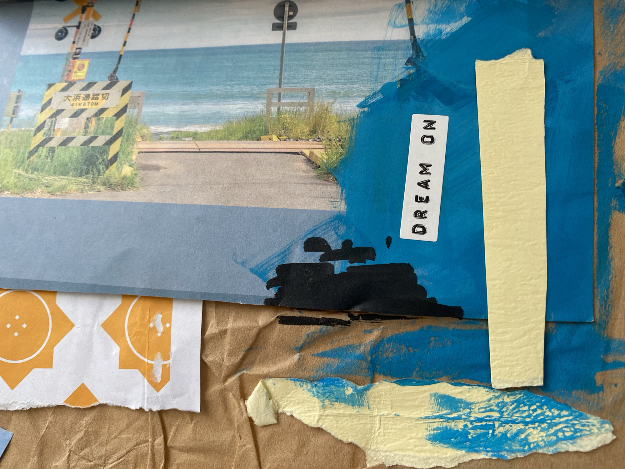 A mixed-media collage. The upper left quadrant features a photograph of a Japanese railroad crossing with a barrier and a sign in Japanese text. The crossing leads to a path towards a body of water, possibly the ocean. The background of the photograph shows a clear blue sky and the ocean. The photograph is attached to a blue piece of paper.
To the right of the photograph and blue paper, there is a large vertical strip of blue paint. Affixed on the blue paint is a white label with the text "DREAM ON" printed vertically. Adjacent to the label is a yellow rectangular piece of paper. Below the blue paper and photograph are pieces of torn paper with an orange geometric pattern, and a piece of painted torn paper with yellow and blue paint. The background of the image is a brown paper with creases.
There is no evidence to suggest that the location is in Lâm Viên - Đà Lạt, Vietnam.