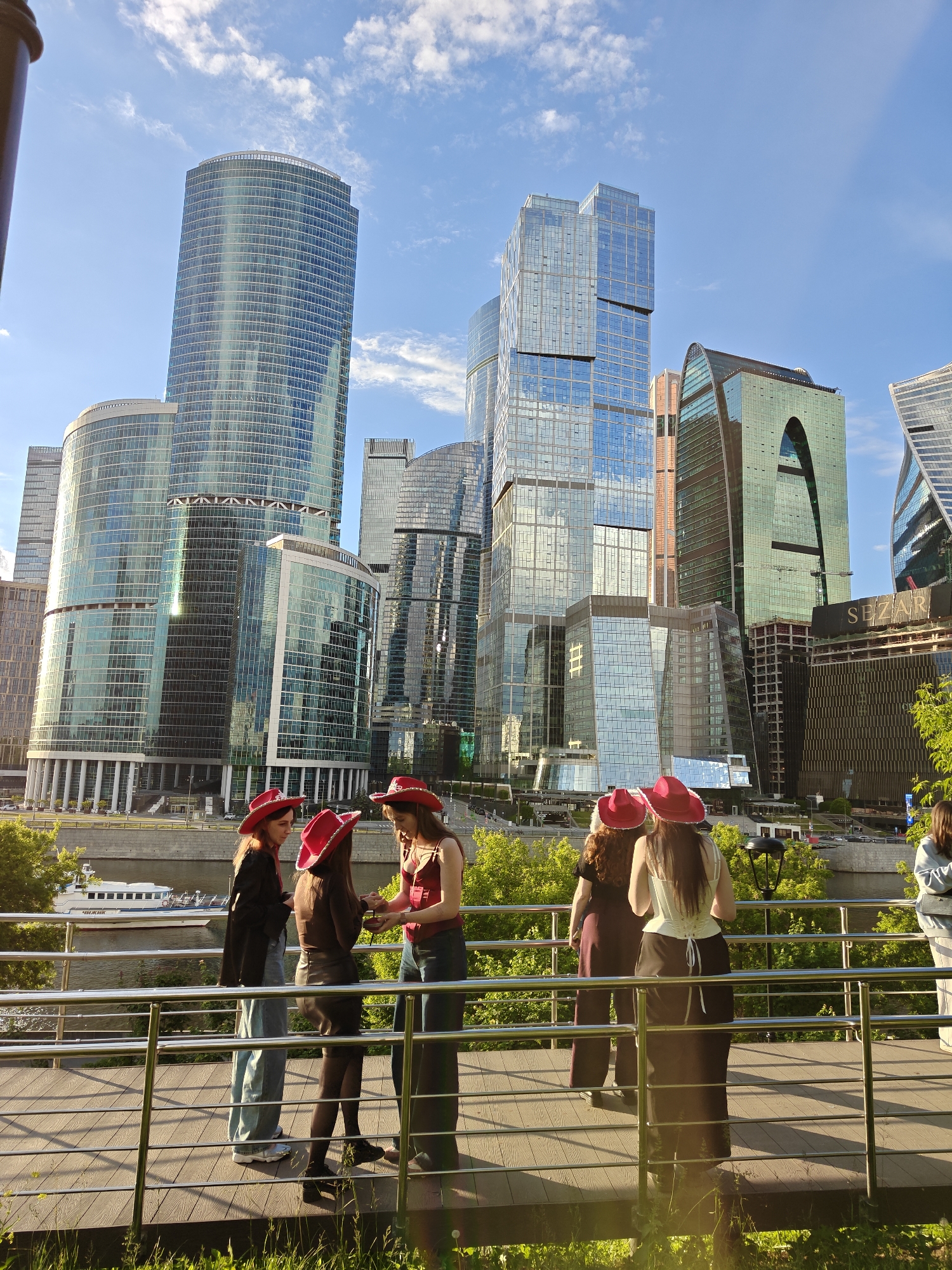 The image depicts a group of six women, all wearing identical bright red cowboy hats, standing on a raised wooden platform with a silver metal railing in front of a cityscape in Moscow, Russia. Behind the platform, a body of water, possibly the Moskva River, is visible, with a white tour boat sailing on it. Lush green trees and vegetation line the riverbank, creating a natural barrier between the water and the urban environment.
The architectural backdrop consists of several modern, high-rise skyscrapers with glass facades, typical of the Moscow International Business Center (Moscow-City). The buildings reflect the blue sky and scattered white clouds above. The "SEZAR" logo is visible on one of the buildings.
The women are dressed in various outfits, including jeans, skirts, blouses, and jackets. They appear to be engaging in conversation. The light suggests it's late afternoon or early evening, as there are long shadows cast on the platform.
