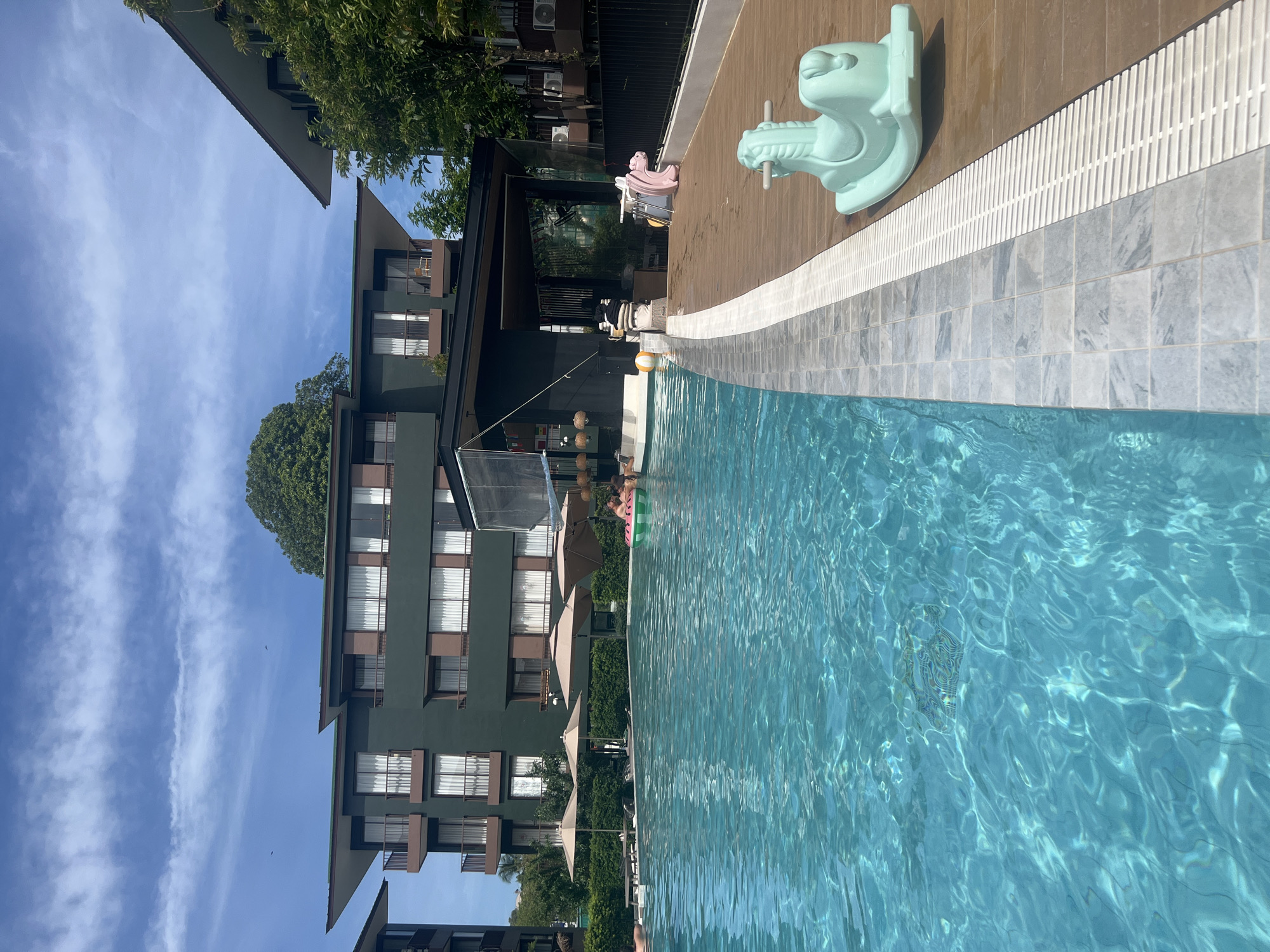 A poolside view at an apartment complex in the city of Ko Samui District, Thailand, under a partly cloudy sky. A bright blue swimming pool occupies the foreground, with its edge lined by grey tiled coping and a white grate. A wooden deck extends to the right of the pool, featuring a mint green rocking horse toy. Beyond the deck and pool, a dark green multi-story apartment building with brown balconies and white-curtained windows is visible. Several beige umbrellas are set up near the building. Two people are partially visible in the pool on the left, and a few more people are in the water on the right, near a green and white inflatable ring. The overall scene suggests a resort or residential area.