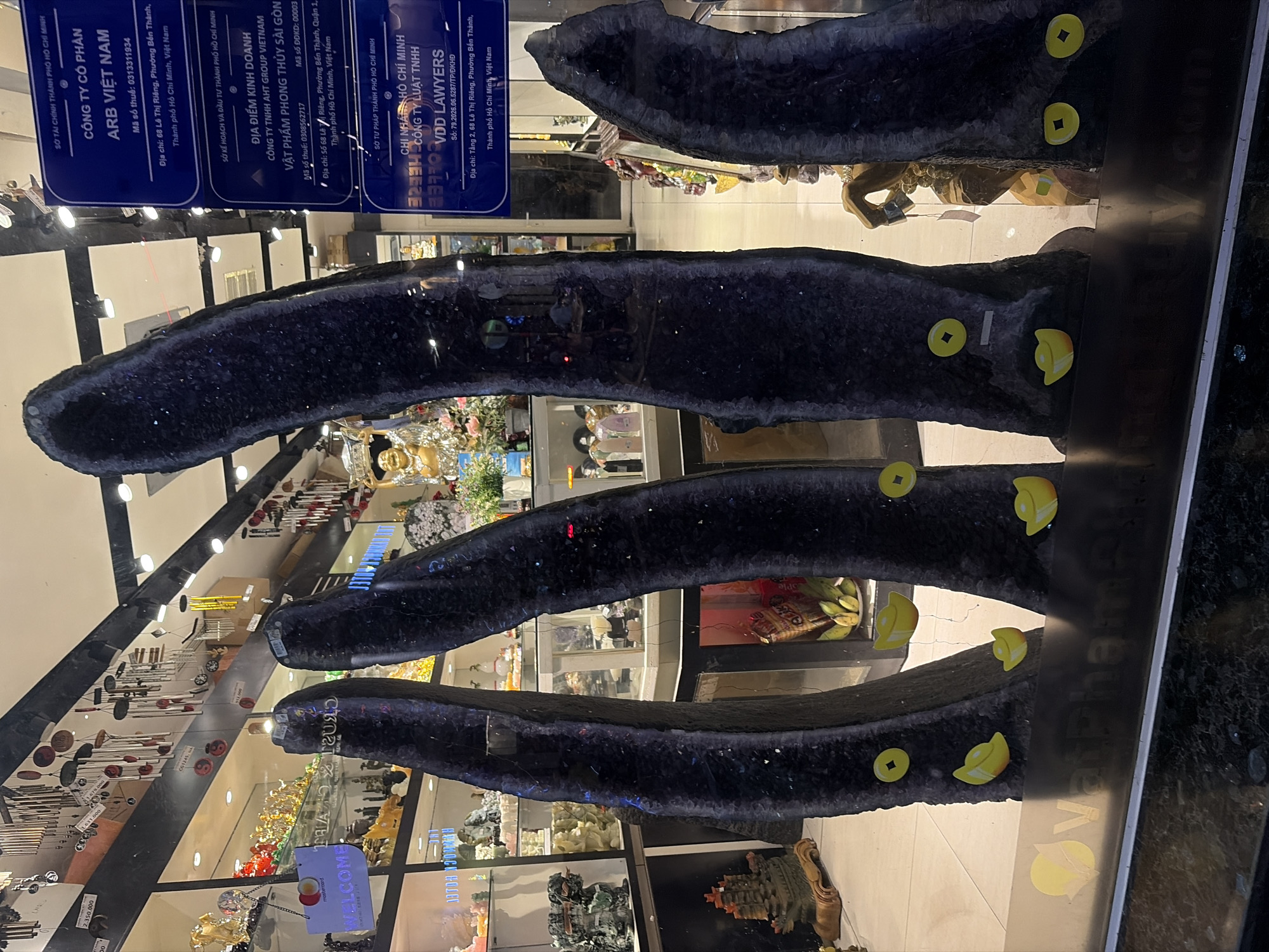 The image shows a storefront display featuring several large, dark purple crystal formations. These crystals are arranged vertically and appear to be natural, uncut specimens. Yellow, coin-shaped decorations and what seem to be ingots are placed at the base of the crystals. Behind the crystals, the interior of the store is visible, showcasing various decorative items, including statues, figurines, and shelves filled with merchandise. A sign on the right side of the image, in Vietnamese, indicates business information, including addresses and company names, one of which is "VDD LAWYERS" located in Thủ Đức, Ho Chi Minh City, Vietnam. The storefront appears to be on a street, as indicated by reflections on the glass.