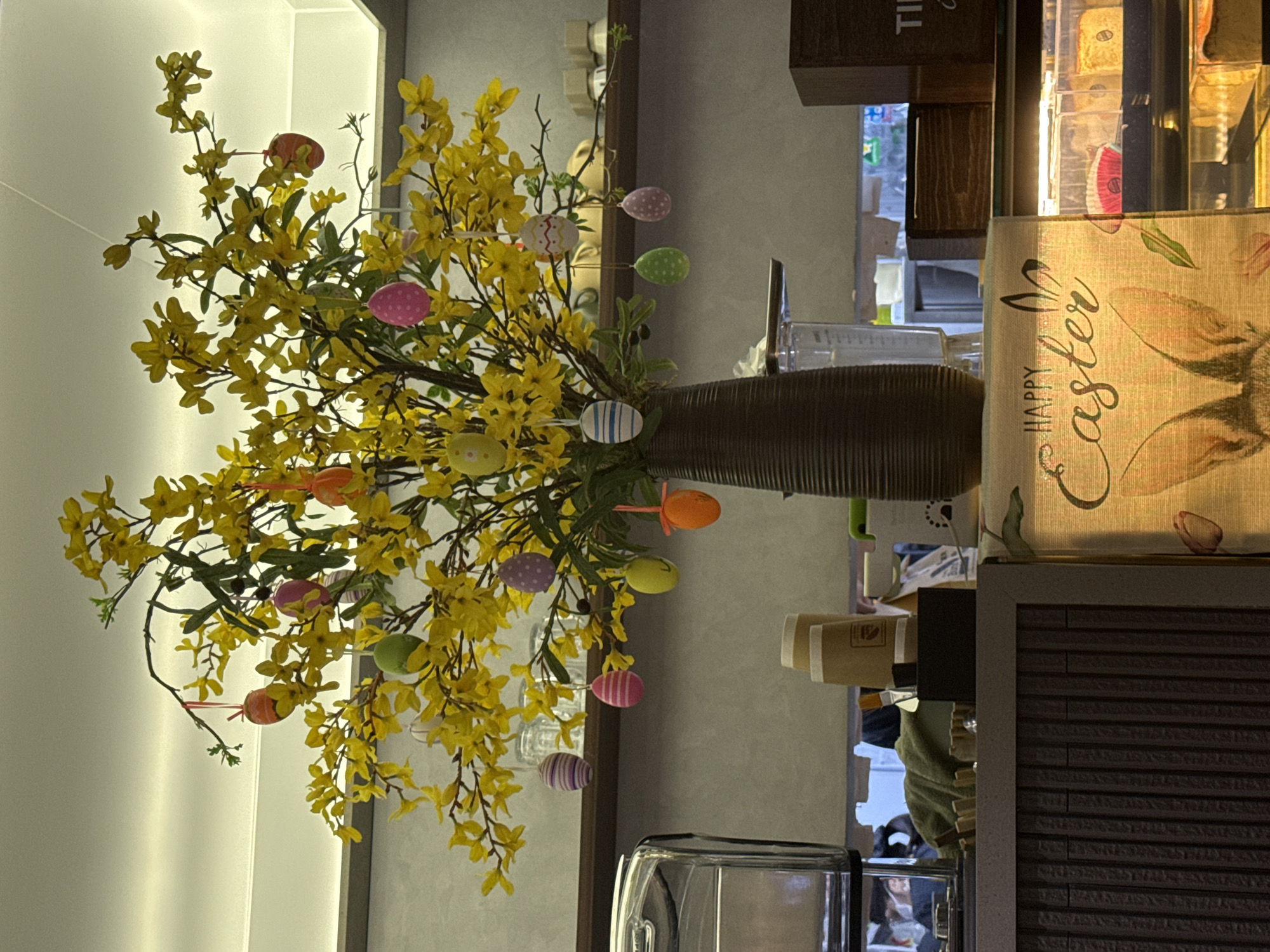 The image captures a vibrant Easter-themed floral arrangement. A large vase filled with yellow flowers adorned with colorful Easter eggs sits prominently in the foreground. The arrangement is placed on a counter or table in what appears to be a cafe or restaurant. Other items in the background include coffee cups, a coffee machine and a 'Happy Easter' decorative lamp with bunny imagery. The background setting seems modern. The photo provides no information regarding Đà Nẵng.