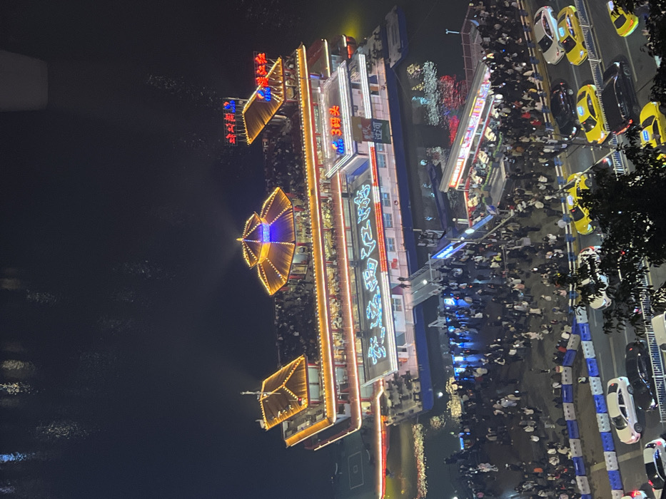 The image depicts a crowded nighttime scene along a riverfront in Yuzhong District, Chongqing, China. A large, brightly illuminated multi-deck vessel, resembling a traditional Chinese pagoda-style ship, is docked at a pier. The vessel features multiple levels with prominent golden-lit curved roofs and extensive string lighting. Neon signage on its hull displays "江海码头" (Jianghai Matou - River-Sea Wharf) in light blue characters, while an upper deck features "3码头" (3 Matou - Pier 3) in similar lighting, and the highest deck displays red neon characters for "斗舰" (Doujian - Warship/Battleship). Numerous individuals populate all visible decks of the vessel.
A well-lit gangway extends from the vessel to a densely packed waterfront area. This area is teeming with people, some gathered near small, brightly lit stalls or structures. Adjacent to the waterfront is a street, partially obscured by foreground foliage, where multiple vehicles, including several yellow taxis and white cars, are observed in motion or parked. Blue and white barriers separate the street from the pedestrian waterfront. The entire scene is viewed from an elevated perspective, looking down onto the illuminated activity against the dark, reflective surface of the river.